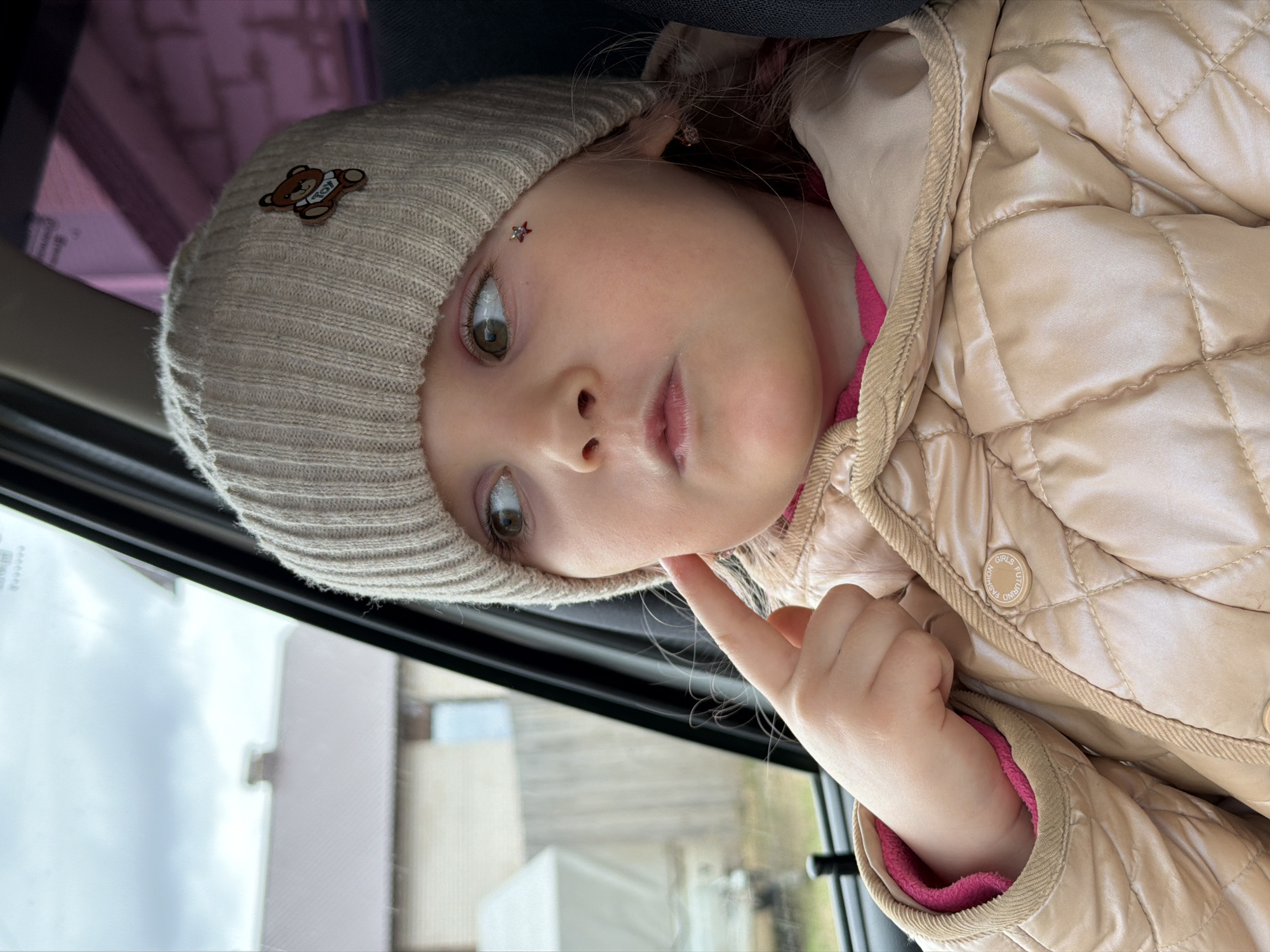 A close-up, portrait-oriented shot, rotated 90 degrees clockwise, features a young child, likely a toddler or preschooler, seated inside a vehicle in Chaikovsky, Russia, looking out a window during the daytime. The child has fair skin and large, expressive greenish-brown eyes, gazing thoughtfully towards the blurry outdoor scene. A small, sparkly red star-shaped jewel or sticker is affixed to their forehead above the left eyebrow, adding a whimsical detail.
The child is warmly dressed in a light beige ribbed knit beanie, adorned with a brown teddy bear patch that has the word "TOY" faintly visible on its chest. They wear a light beige quilted jacket, which appears shiny and padded, with a bright pink inner lining peeking out at the collar and sleeve cuffs. A circular button on the jacket clearly displays the words "FASHION GIRLS FUTURE". One of the child's fingers gently rests on their chin, suggesting curiosity or contemplation.
The scene outside the vehicle's window is largely out of focus, showing indistinct light-colored buildings or structures against a light sky, indicative of an urban or suburban environment. The presence of the window frame and the blur outside confirms the child is inside a moving or stationary car. The overall impression is one of a quiet, reflective moment captured during travel.