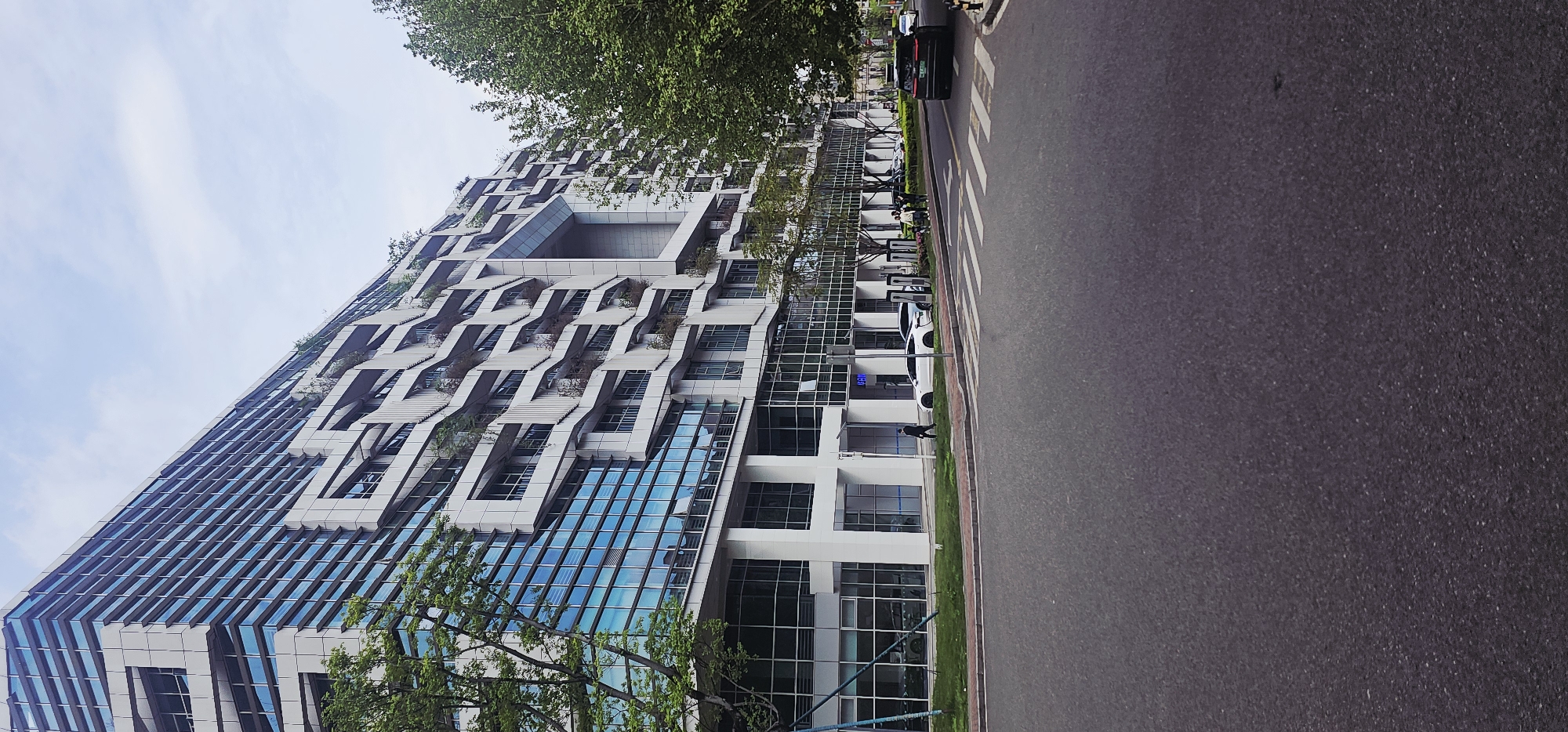 The image shows a modern building in Jiulongpo District, China. The building features a glass facade on the lower floors, transitioning to a unique, terraced design with a central square opening on the upper floors. The terraces appear to have some vegetation planted on them. A road runs along the side of the building with a crosswalk visible. A black car is driving on the road. On the sidewalk along the building, charging stations are placed, and one person wearing a mask is walking. Trees are also located beside the building. The sky is overcast.