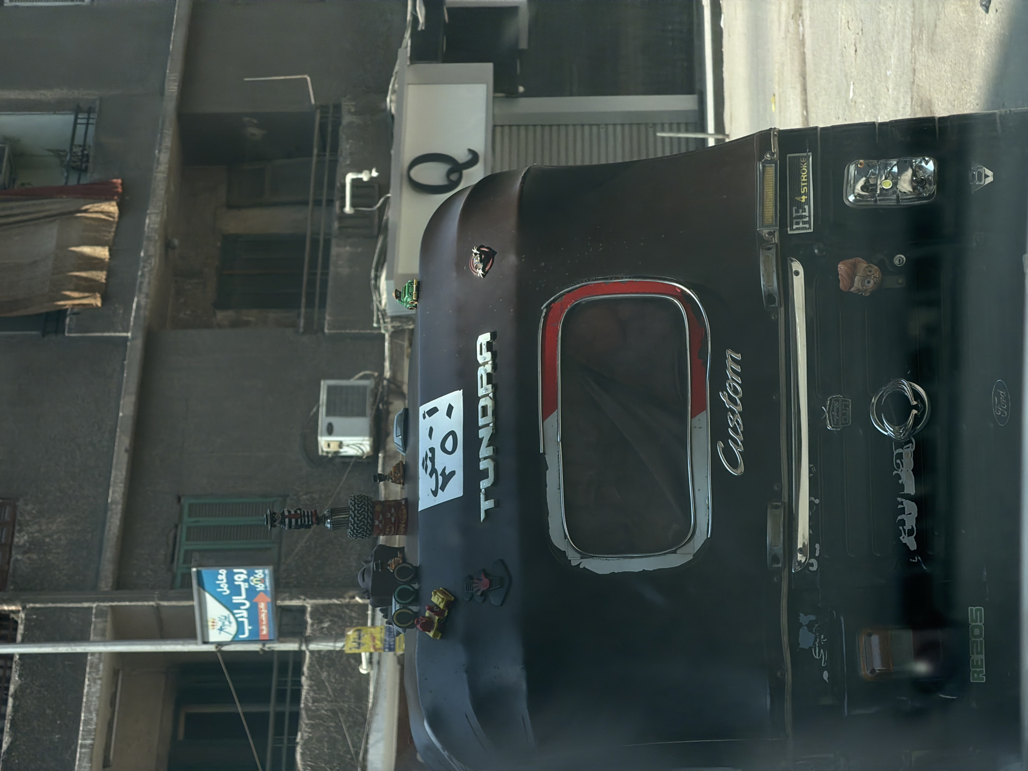 In Cairo, Egypt, this photograph captures the rear of a customized three-wheeled vehicle, commonly known as a tuk-tuk. The vehicle is dark brown and adorned with various decorative items. On the roof, a small green toy car and a miniature yellow vehicle are placed. A white license plate with Arabic script and numbers is attached to the back, along with the word "TUNDRA" in silver lettering. Further down, the word "Custom" is inscribed in silver script. Various stickers and emblems are also visible on the vehicle's exterior, including a small face sticker and a "HE 4 STROKE" badge. The backdrop features a building with concrete walls and windows, characteristic of urban architecture in Cairo. A street sign with Arabic writing is mounted on a pole to the left. The overall scene depicts a typical street view in Cairo, highlighting the unique personalization of vehicles in the city.