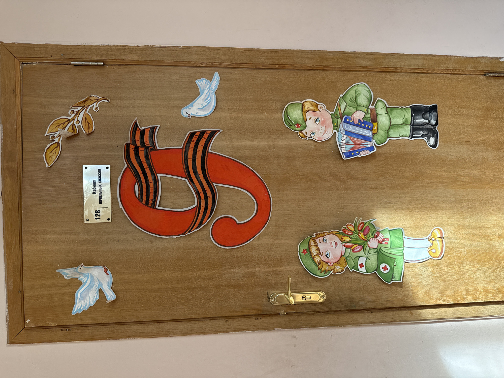 The image shows a wooden door, likely to a classroom given the sign on the door that says "Cabinet of Elementary Classes 128." The door is decorated with symbols associated with Victory Day, a Russian holiday commemorating the victory over Nazi Germany in World War II.
Symbols on the door include:
* An orange ribbon with black stripes, known as the Ribbon of Saint George, a military symbol.
* Two paper cutouts of white doves.
* A branch with leaves.
* A drawing of a male soldier playing an accordion.
* A drawing of a female medic holding tulips.
The door is light brown, and the background is a light pink wall. There is a brass door handle on the door. The location in the city of Rusanovo, Russia is only specified by implication based on the Victory Day decorations, since the city is not explicitly mentioned in the image.
