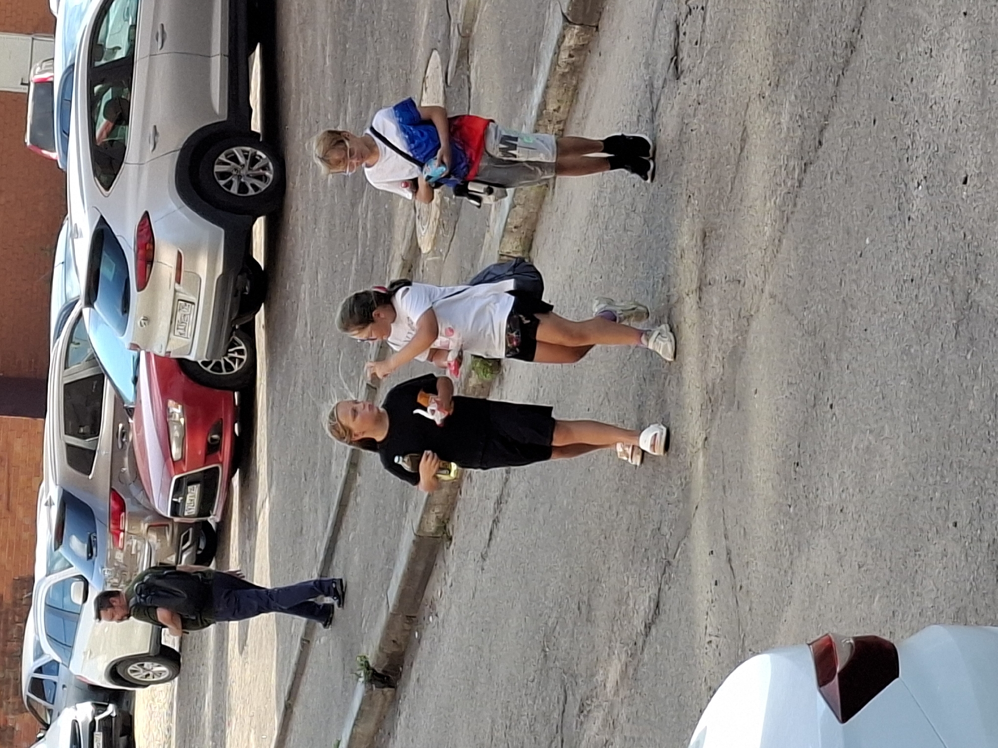A man walks with a black backpack on his back in a city street, in the background several cars are parked. On the sidewalk three young people walk. Two girls and a boy. The girl on the left wears a black t-shirt and black shorts, holding a bottle in her right hand and a snack in her left hand. The middle girl wears a white t-shirt and dark shorts, she is holding a bottle in her left hand. The boy on the right wears a white t-shirt with blue and red accents, gray shorts, and black shoes. He has a bag over his right shoulder. The street is paved and has a sidewalk. The background shows buildings and parked cars.