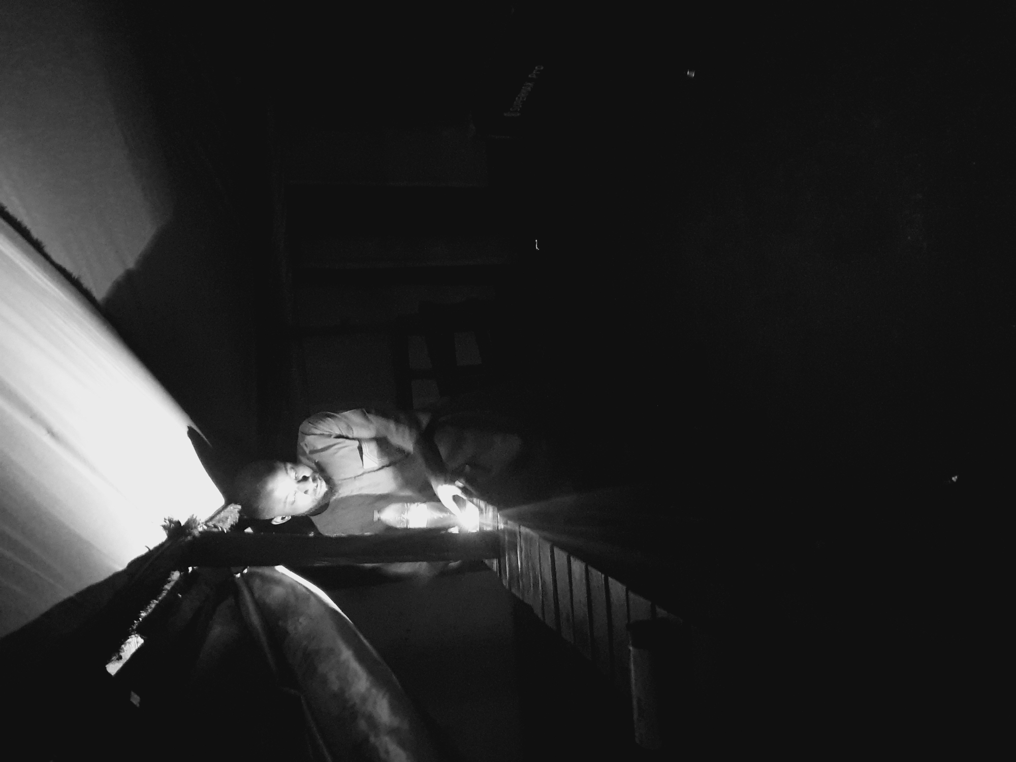A medium shot in black and white captures a young man in a dimly lit, rustic structure. He is seated on a wooden platform, leaning forward with his head down, illuminated by a light source that appears to be a bottle of water held in his hand. The light casts a strong glow on his face and the surrounding area, creating harsh shadows. He is wearing a collared shirt. The structure seems to be covered by a corrugated roof or tarp, with wooden supports visible. The overall atmosphere is one of confinement and a possibly makeshift setting. The image suggests a moment of quiet contemplation or work under limited lighting conditions. Without further context, pinpointing a specific location within Lokoja, Nigeria, is not possible based solely on the visual information presented.