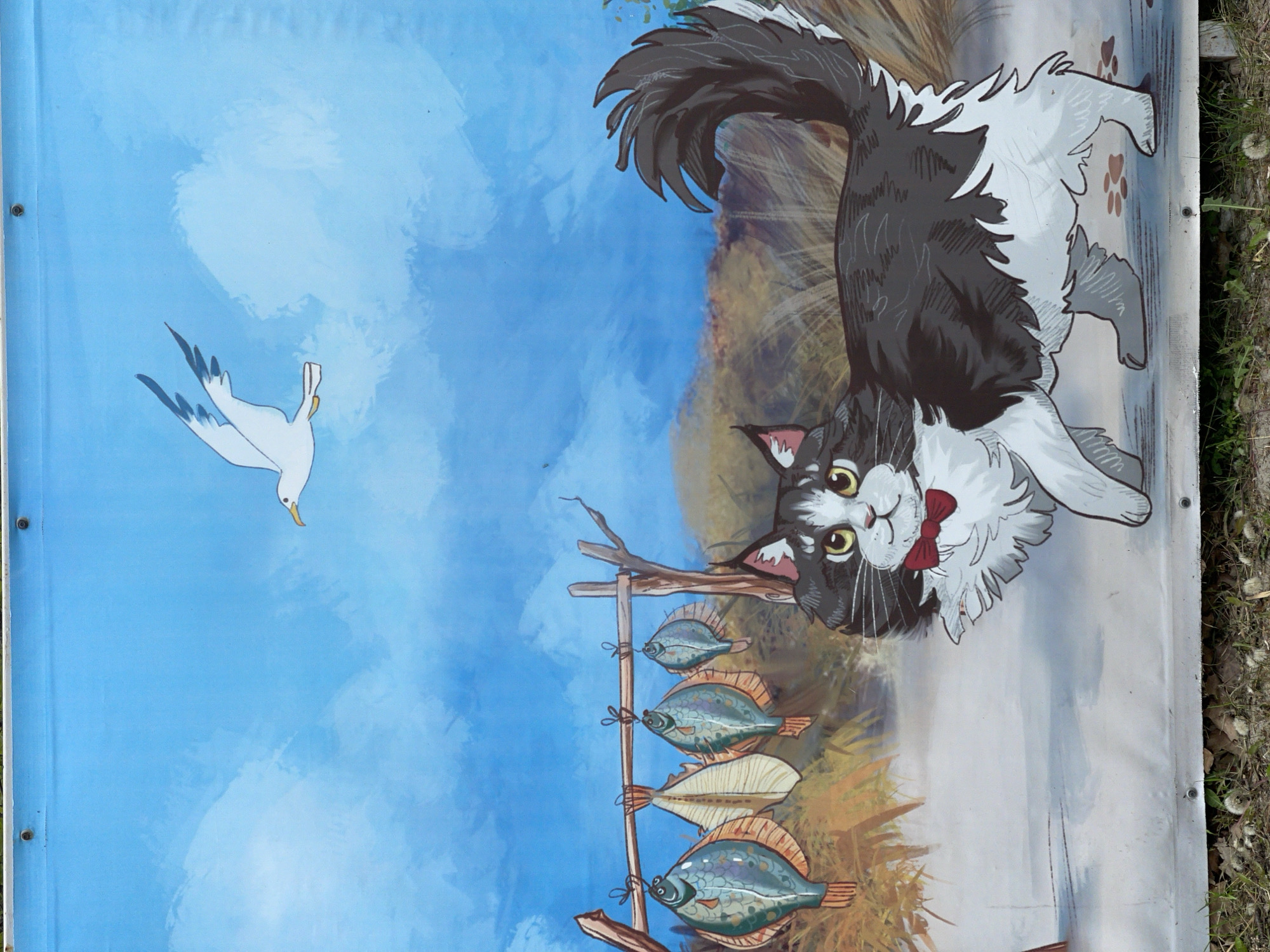 The image displays a vibrant, cartoon-style illustration on what appears to be a banner or mural. The main subject is a fluffy black and white cat, depicted with a white chest, paws, and face, and a dark grey/black body and tail. It wears a red bow tie and has large, attentive yellow eyes. The cat is positioned on the right side of the scene, appearing to look upwards and to the left.
In the upper-left portion of the image, a white seagull with a yellow beak is flying against a light blue sky with wispy white cloud-like patches. Below the seagull and extending towards the center of the scene is a simple wooden structure, resembling a drying rack, from which four flatfish (resembling flounder) are hanging by strings. The fish display varying patterns and colors.
The background is divided into a clear blue sky in the upper half and a brownish-green grassy or earthy landscape with some faint vegetation in the lower half, situated behind the cat and the fish rack. Brown paw prints are visible on the ground near the right edge of the banner. The overall scene suggests a coastal or lakeside setting, fitting for Zelenogradsk, a town known for its cat theme. The illustration has a playful and whimsical tone, depicting a curious cat observing its surroundings.
There is no visible text in the image.