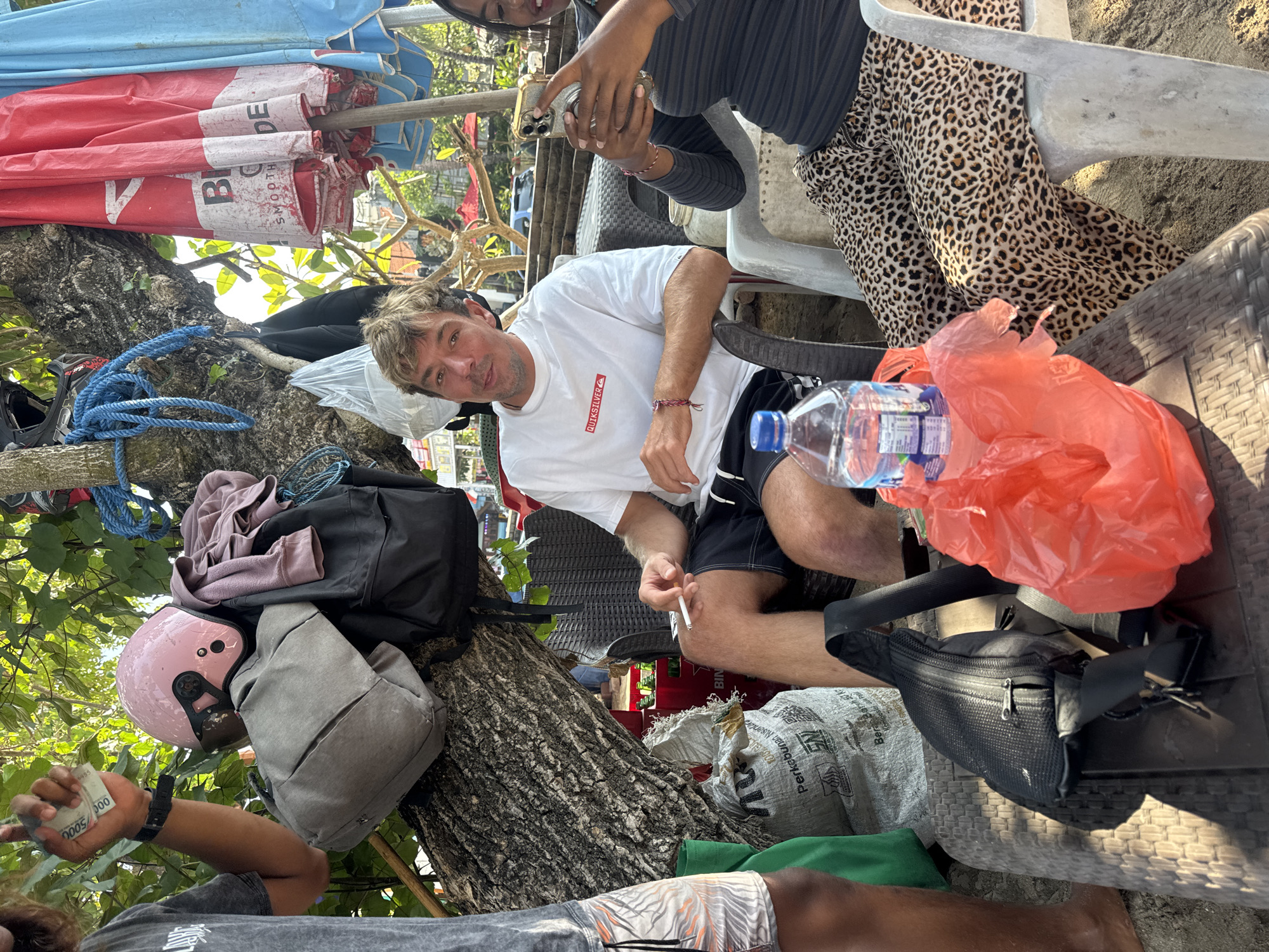 The image depicts a scene outdoors, likely on a beach in Kuta, Indonesia. In the foreground, a man in a white Quiksilver t-shirt and black shorts is seated, holding a cigarette. To his right, a woman wearing a dark striped top and leopard-print pants is holding a smartphone, appearing to take a photo. In front of them, on a textured table, are a plastic water bottle and a bright orange plastic bag.
Behind the seated individuals, a tree serves as a backdrop, with several items hanging from its branches. These include a pink helmet, a grey backpack, a black backpack, and a blue rope. A person's arm is visible on the left, holding several Indonesian Rupiah banknotes. A large woven bag with Indonesian text is also present near the tree trunk. In the background, beach umbrellas and a wicker seating area are visible, suggesting a commercial establishment or relaxation spot. The ground appears to be sandy, consistent with a beach environment.