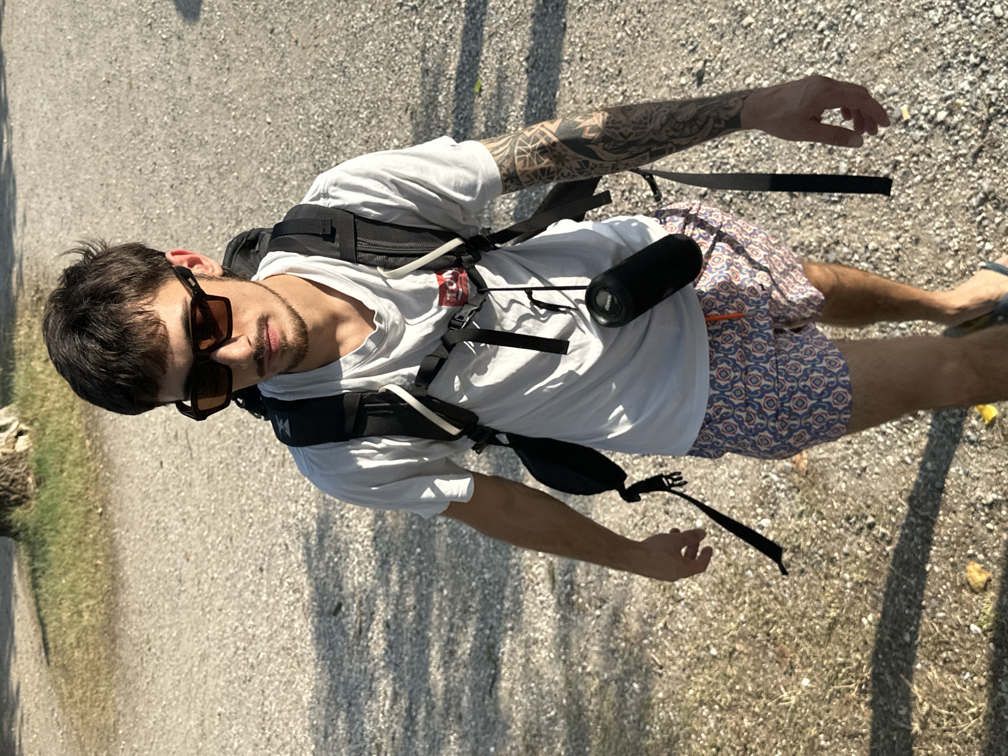 A young man with dark hair and a goatee is captured in a medium, high-angle shot, walking outdoors. He is wearing a white t-shirt, patterned swim trunks, and sunglasses. A black backpack with straps is visible on his back, and a black cylindrical object, possibly a portable speaker, is attached to the backpack. His left arm, tattooed with intricate black designs, is partially visible. The man appears to be walking on a gravel path, with patches of green grass to his left and shadows cast by trees on the ground to his right, suggesting a sunny day. The image is taken in portrait orientation, with the subject positioned slightly off-center to the left. There is a small red and white tag visible on the chest strap of his backpack. The overall impression is of a casual, outdoor excursion.