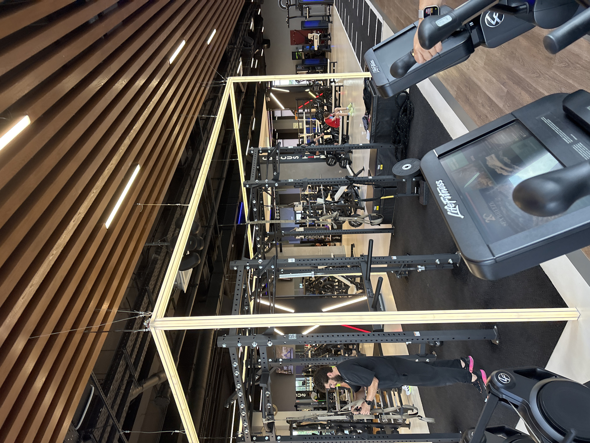 The image depicts the interior of a modern, well-lit gym. A man wearing a black t-shirt and black pants, with pink athletic shoes, is standing near a weight rack, holding a piece of equipment. In the foreground, two exercise machines with digital displays are visible, one with a hand on the handlebars. Other gym equipment, including weight racks, benches, and cardio machines, are arranged throughout the space. The gym features a wooden slat ceiling with recessed lighting and exposed industrial elements. The flooring consists of black rubber mats in the workout areas and a lighter colored flooring elsewhere. The overall environment suggests a professional and well-maintained fitness facility. This scene is located within the city of Monolit, Russia.