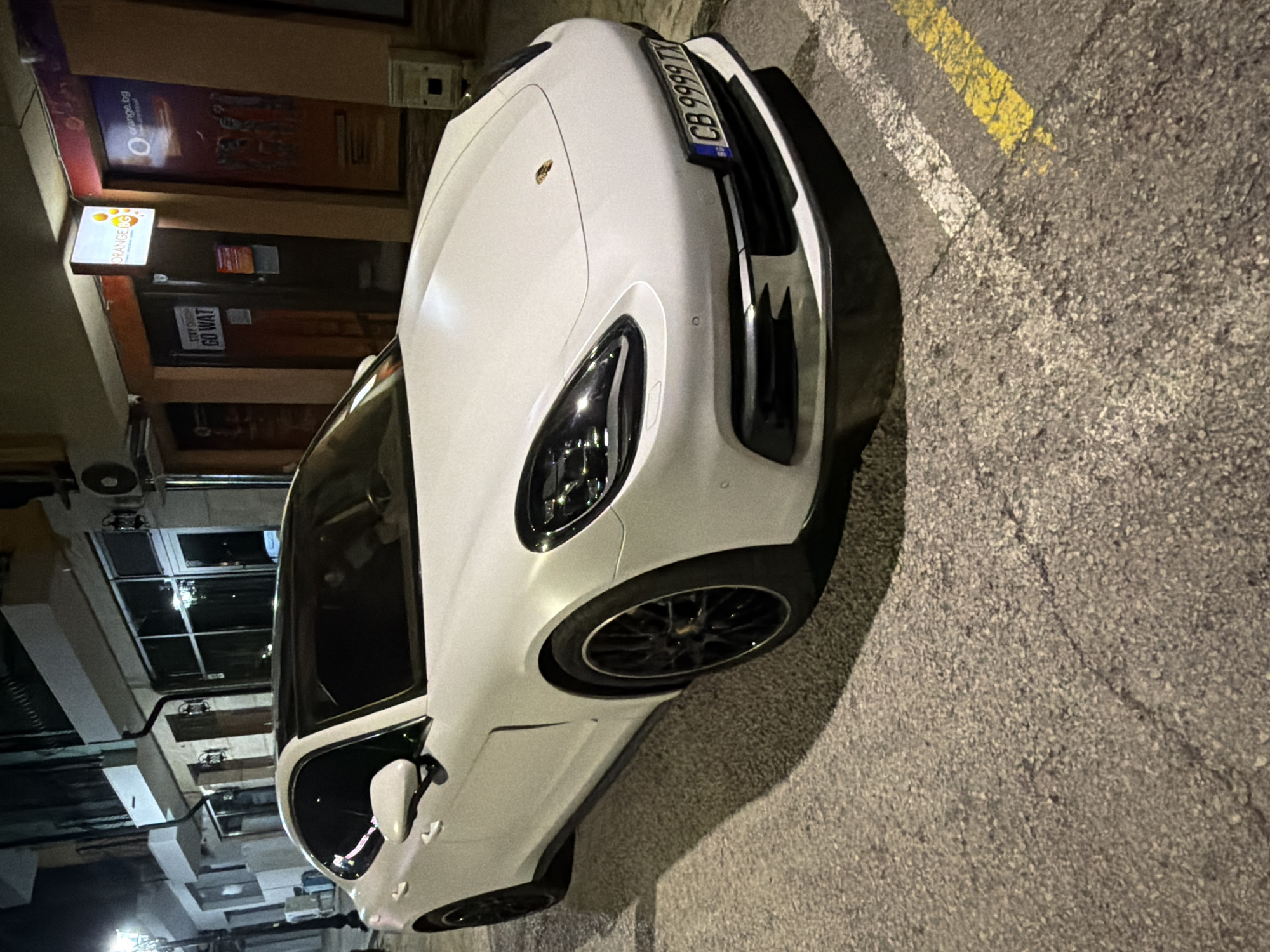 A white Porsche Panamera is parked on a city street at night in Veliko Tarnovo, Bulgaria. The car is facing right, with its front bumper and license plate clearly visible. The license plate reads "CB 9999 TX". The car is parked on asphalt, with yellow and white painted lines indicating a parking space. In the background, there are buildings with shopfronts and illuminated signs. One sign reads "ORANGE" with the Orange logo. The street is illuminated by streetlights and lights from the buildings. The overall scene suggests an urban environment at night.