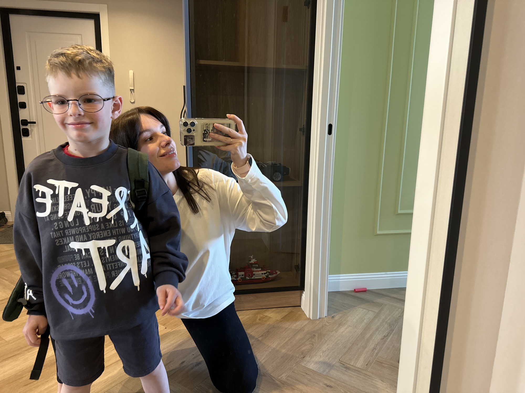 A woman is taking a photo of a young boy in front of a mirror. The boy is wearing glasses, a gray shirt, and black shorts with a backpack. The woman is kneeling and holding her phone up to take the picture. In the background, there is a door, a cabinet, and a wall. There appears to be a toy fireboat in the cabinet. The floor is wood. The location could be a home in Chaikovsky, Russia.