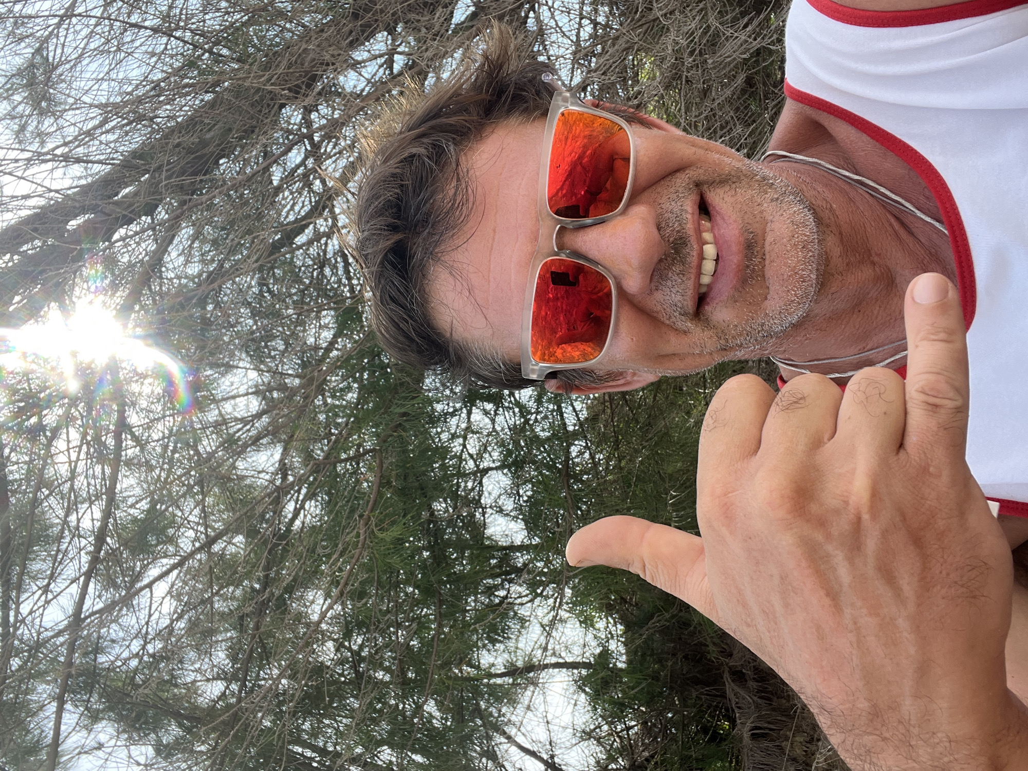 A man, wearing sunglasses with red reflective lenses and a white tank top with red trim, is pictured outdoors. He is smiling and giving a "shaka" hand gesture with his left hand. His right hand is giving a thumbs-up. The background consists of dense, dry tree branches and foliage, with the sun shining brightly through the trees, creating lens flare. The setting is likely Mui Ne, Vietnam, given the dry, sparse vegetation consistent with the coastal arid climate of the region.