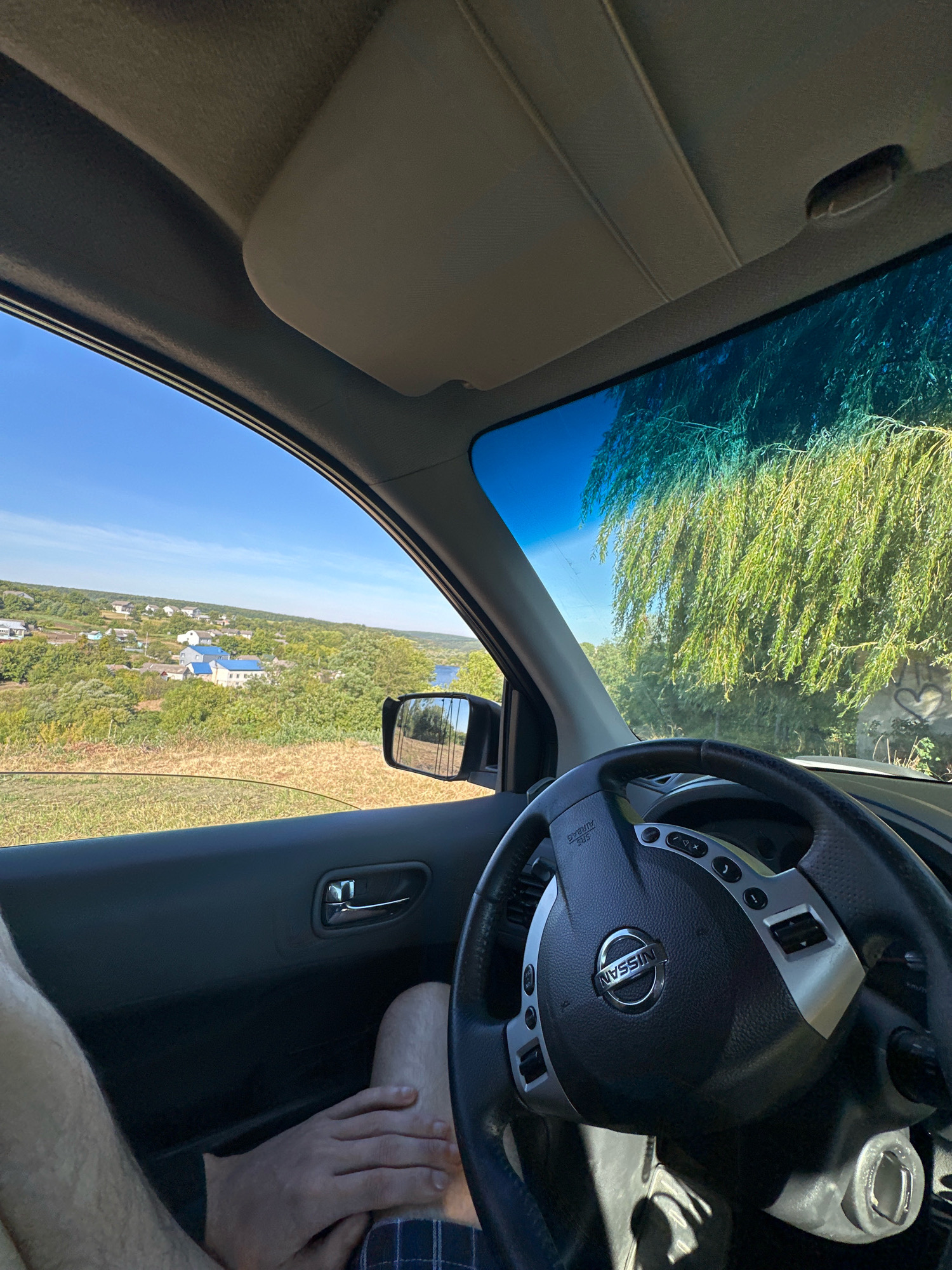 The image shows the interior of a Nissan vehicle, viewed from the perspective of the driver. The steering wheel, with the Nissan logo prominently displayed, is in the foreground. The dashboard and a portion of the passenger seat are also visible. Through the side window, a rural landscape unfolds, featuring rolling hills covered in lush green vegetation. A small village with houses, some with blue roofs, is nestled in the valley. A body of water, likely a river or lake, can be seen in the distance. A large weeping willow tree with long, flowing branches is visible outside the windshield, partially obscuring the view of the sky. The sky is a clear blue, suggesting a sunny day. The car appears to be parked or moving slowly along a dirt road. The overall impression is one of a scenic drive through the Ukrainian countryside, possibly near Makiv, given the rural setting and architectural styles.