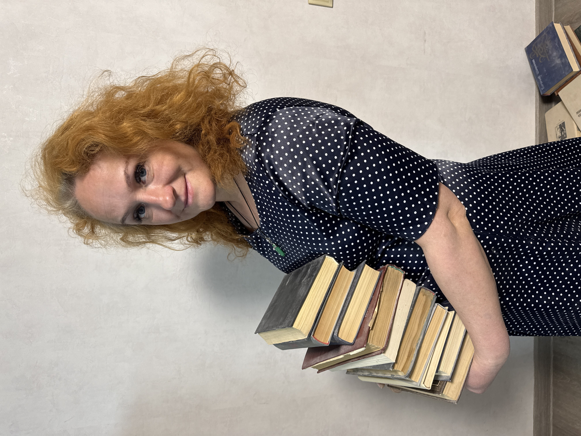 The original image, presented rotated 90 degrees clockwise, depicts a woman indoors. She has voluminous, curly red hair and blue eyes, and is looking towards the viewer with a gentle smile. She is wearing a dark blue dress adorned with small white polka dots and a small green pendant on a dark string around her neck. The woman is holding a stack of approximately eight to ten old, worn books in her arms. The background consists of a plain, light-colored wall. To the right, a few more books are stacked on a darker surface, one of which is a blue book with Cyrillic text on its spine, partially visible as "Европа и Цыганская" (Europe and Gypsy). The setting appears to be a casual indoor environment, possibly a home or study, conveying a studious or relaxed atmosphere. The location context is Chaikovsky, Russia.