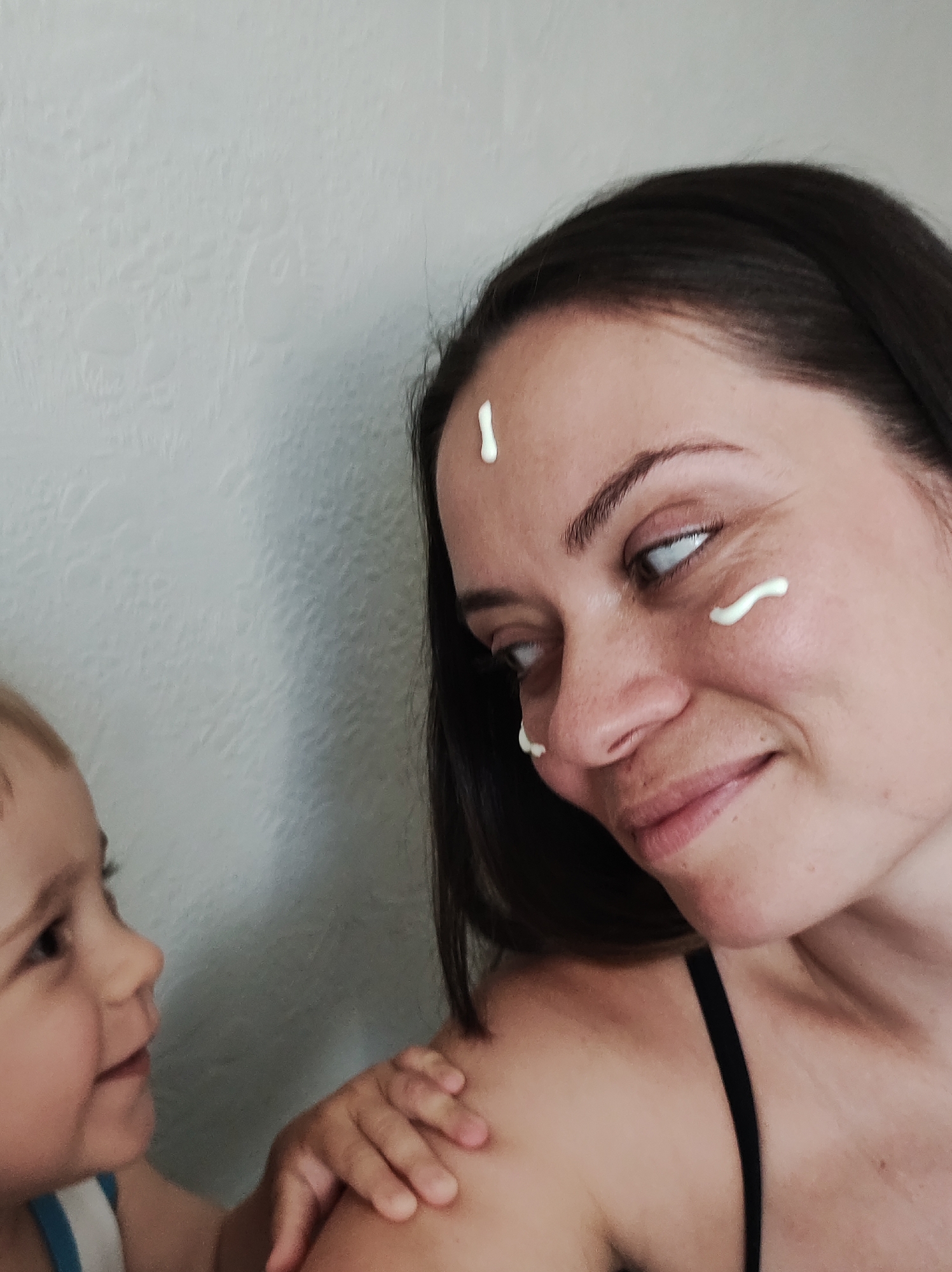A woman with dark hair and fair skin is posing for a photo with a young child. The woman has three dollops of white cream on her face – one on her forehead, one under her eye, and one near her nostril. She is looking toward the child and smiling slightly. The child, who appears to be a toddler, has light hair and skin, and is gazing up at the woman. The child's hand is resting on the woman's shoulder. The woman is wearing a black tank top. The background is a textured, light-colored wall. The location is Chaikovsky, Russia.