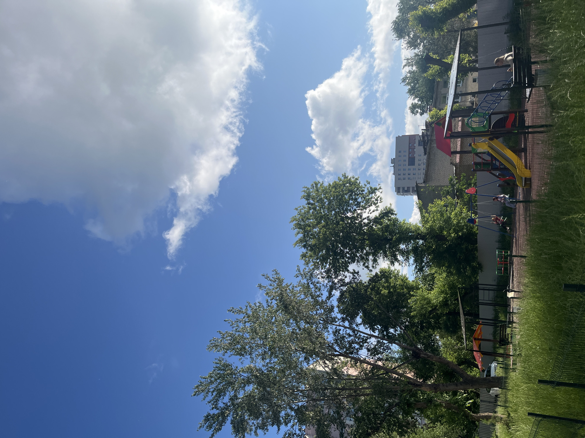 A bright, daytime outdoor scene depicts a children's playground in Chelyabinsk, Russia. The playground, featuring a yellow slide, swings, and climbing structures, is situated in front of a fence and surrounded by lush green grass and trees. In the background, buildings and apartment complexes are visible, indicating an urban setting. The sky is a vibrant blue, with scattered white clouds. A few people are present in the playground, including one woman on a swing and two children on another swing. The overall impression is one of a sunny day in a community park.