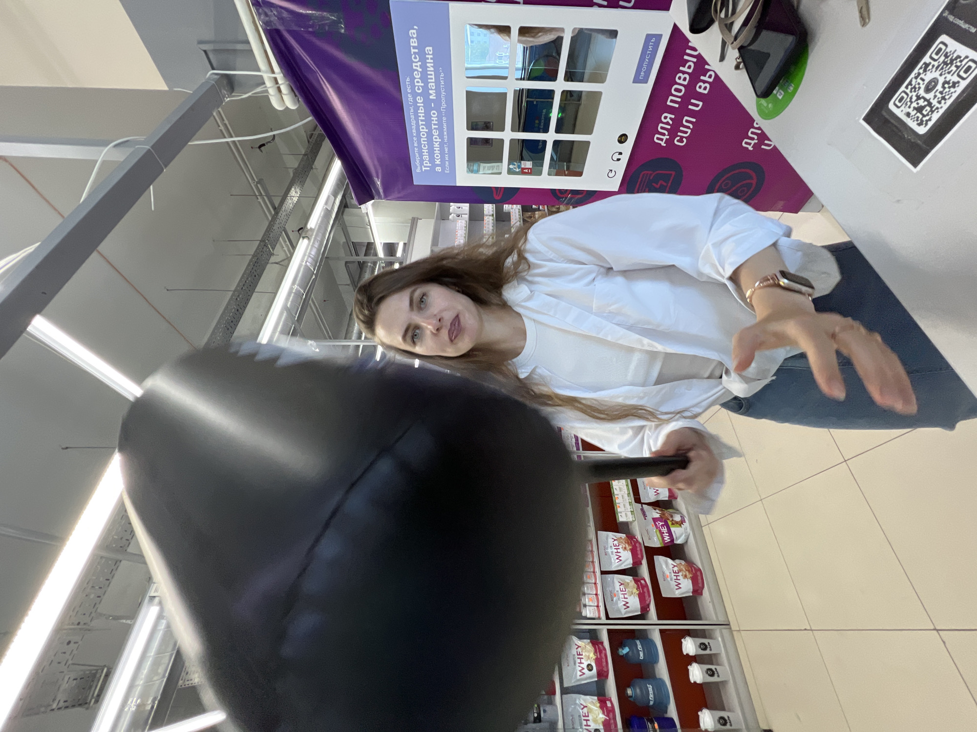 A woman stands in a store, possibly a pharmacy or supplement shop, holding a large, black inflatable object that obscures part of her body and the background. She is wearing a white unbuttoned shirt over a white top and blue jeans. Her left hand is extended forward, and she appears to be interacting with the inflatable object. To her right, there is a purple display with a screen showing a grid of images and text in Russian. The shelves behind her are stocked with various products, including bags labeled "WHEY" and large water bottles. The floor is tiled with beige squares. The ceiling has fluorescent lighting and visible ventilation systems.