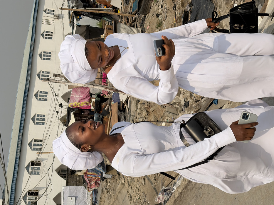 The image portrays two young adult females walking on a rough, unpaved surface in Igboefon, Nigeria. Both women are dressed in matching white, long-sleeved garments—either dresses or jumpsuits—and distinct white, voluminous head coverings.
The woman on the left, positioned slightly ahead, exhibits a soft smile, gazing towards the right side of the frame. She wears small gold hoop earrings and a thin gold necklace. A black shoulder bag with a metallic clasp is slung across her body, and she holds a light-colored smartphone in her right hand.
The woman on the right is depicted looking downwards at a dark-colored smartphone held in her right hand, displaying a subtle smile. She also wears a thin gold necklace and carries a black handbag with a strap and buckle design in her left hand.
The background features a large, multi-story white building with a blue roofline and several arched windows, along with an external air conditioning unit. Overhead, a tangle of electrical wires is visible. The immediate surrounding environment is characterized by an uneven, debris-strewn ground comprising dirt, rubble, wooden planks, and various discarded materials. Improvised structures, including what appears to be a market stall with a partially visible pink banner displaying "St. Gabriel," are present in the midground. The scene is illuminated by bright daylight.