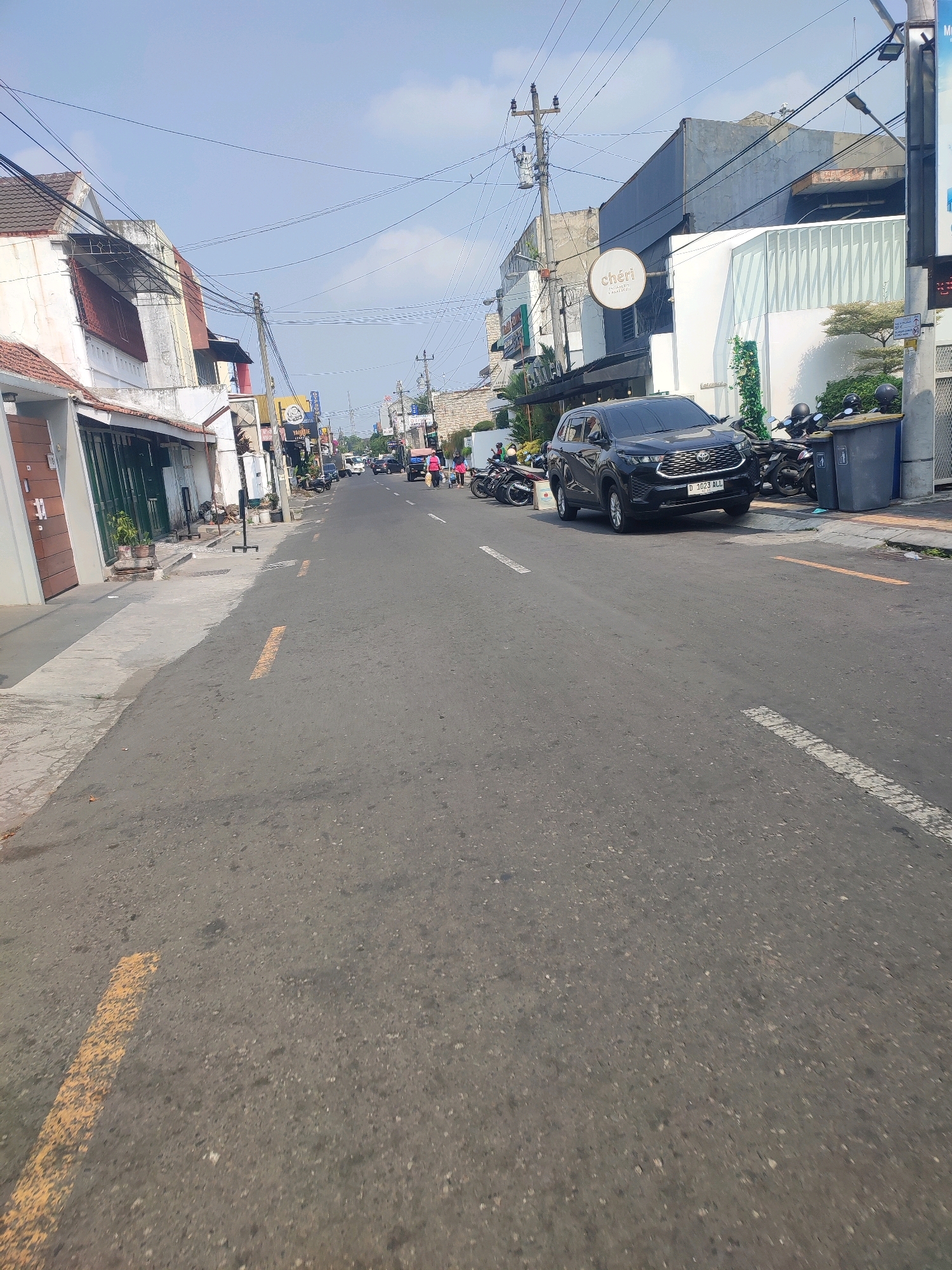 The image depicts a street scene in Yogyakarta, Indonesia, likely in an urban commercial area. A black SUV with license plate D 3023 ALL is parked on the right side of the road, facing away from the camera. Next to the SUV are several motorcycles and a large gray garbage bin. Across the street, there are buildings with commercial signage, including one with the logo "chéri". The street is paved and has painted yellow and white lines indicating traffic lanes. The sky is clear with some scattered clouds. There are a few pedestrians visible in the distance, including two individuals walking on the right side of the road. Power lines and utility poles are present throughout the scene, indicating the urban setting. The architecture of the buildings suggests a blend of modern and older styles.