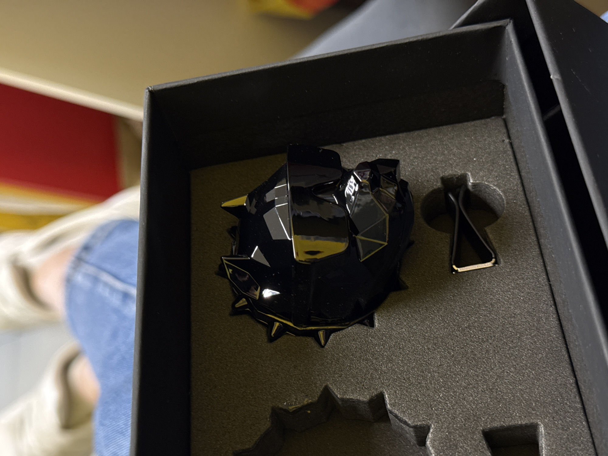 A close-up shot shows a black, spiky air freshener inside a black box. Next to the air freshener is a black car vent clip with a silver metal tip. The box is lined with a gray foam material. The scene appears to be indoors. Part of a person is visible in the background wearing jeans and a light-colored shoe. The location is Unknown, Unknown.