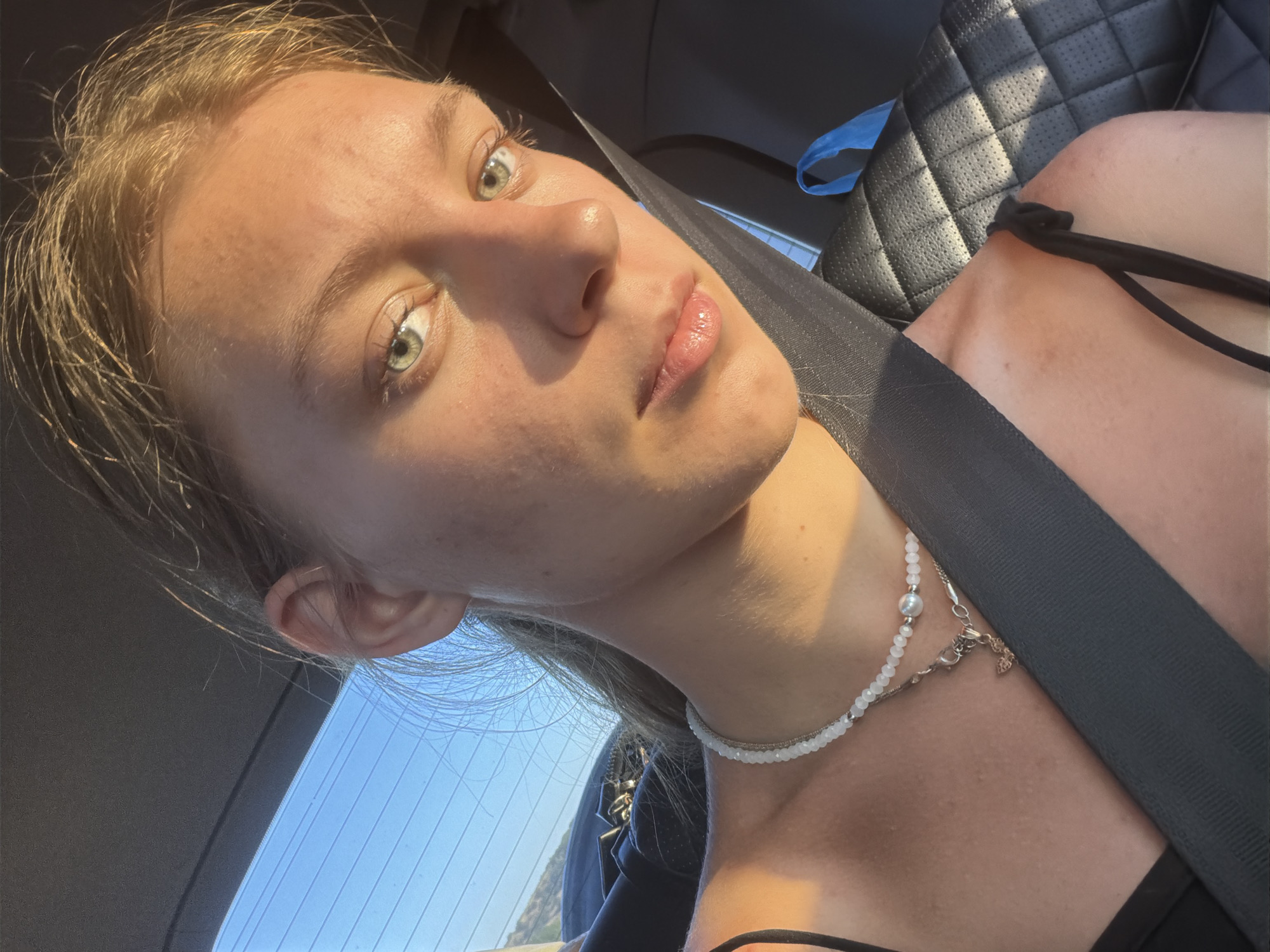 A young Caucasian female with blonde hair pulled back is captured in a selfie taken from inside a car, illuminated by the golden hour sunlight. She is wearing a black top and a white beaded necklace with a silver pendant. A seatbelt is visible across her chest, and the background shows a car's interior with black quilted seats and a blue bag. The overall impression is one of a casual, sun-drenched moment during travel. The image does not provide any visual cues that would allow for the identification of the location as Belogorki, Russia.