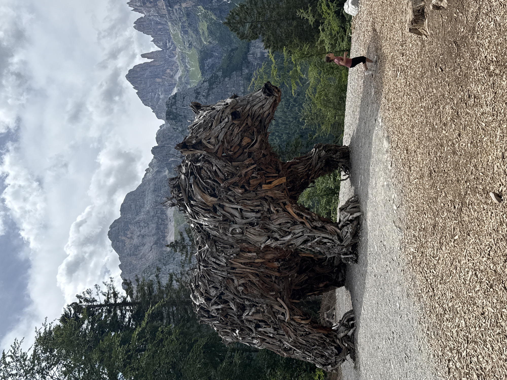 The image depicts a large, intricately constructed bear sculpture made of driftwood, positioned outdoors in an environment characterized by natural elements. The sculpture is located in Molveno, Italy, specifically at the Forest Bear Park. In the background, towering, rugged mountains with rocky peaks dominate the skyline under a cloudy sky. Lush green trees and forests surround the sculpture, suggesting a mountainous, wooded area. In the foreground, a young girl wearing a pink shirt and black leggings is captured in mid-stride, running across a ground covered with wood chips and gravel. The bear sculpture is the central and largest object, its form rendered with numerous pieces of weathered wood. The scene conveys a sense of natural beauty and artistic installation within a striking mountainous landscape.