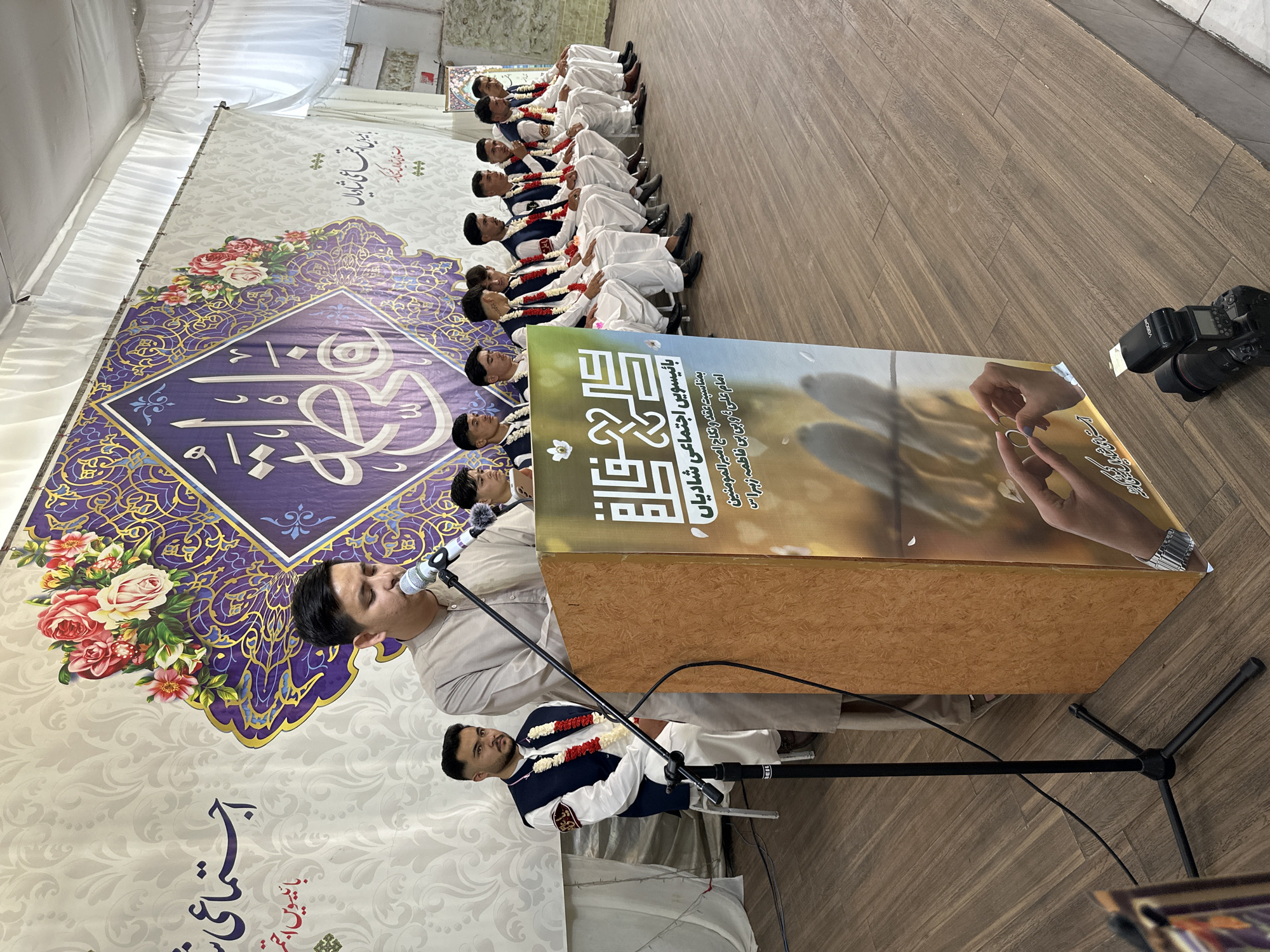 Here is a detailed description of the image:
**General Scene:** The image captures an indoor event, likely a wedding ceremony, featuring a group of men in traditional attire. The scene is set within a tent-like structure with ornate banners and decorations.
**People:** A line of men in matching white outfits with dark blue vests and floral garlands are seated. Another man in a similar outfit stands at a microphone stand, addressing the audience. Another man can be seen with a beard and similar attire in the foreground.
**Objects:**
* **Banners:** A large banner behind the seated men features a floral design and Arabic script. A smaller banner is positioned near the speaker, showcasing a graphic of hands exchanging a wedding ring.
* **Microphone Stand:** A black microphone stand holds a microphone used by the speaker.
* **Camera:** A digital camera with a flash unit is seen on the floor.
**Actions:** The seated men appear to be attentively listening to the speaker. The speaker is actively addressing the audience through the microphone.
**Interactions:** The speaker interacts with the audience through his address. The men in the line seem to be interacting by listening to the event.
**Environment:** The location appears to be a tent-like structure, possibly a temporary setup for the event. The floor is made of wood-like material.
**Text on Banners:** The banner near the speaker has a graphic of a ring being placed on a finger, it has Arabic text.