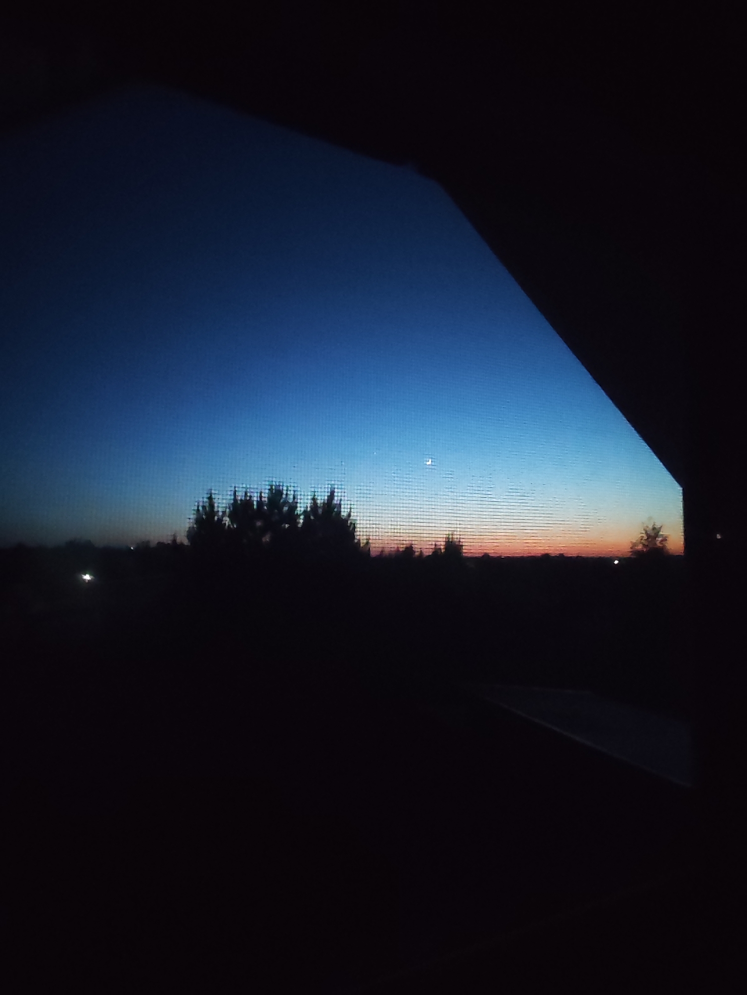 The image shows a view from inside a building, likely a window or balcony, looking out onto a dark landscape. The sky is a gradient of color, transitioning from a dark blue at the top to orange and pink hues near the horizon. There is a small, faint crescent moon visible in the upper portion of the sky. The horizon is populated with silhouetted trees and possibly distant buildings, creating an undefined dark mass. A portion of the building's structure frames the scene, obscuring parts of the view on the right and top. The location of the image is Фоки, Russia is unknown.