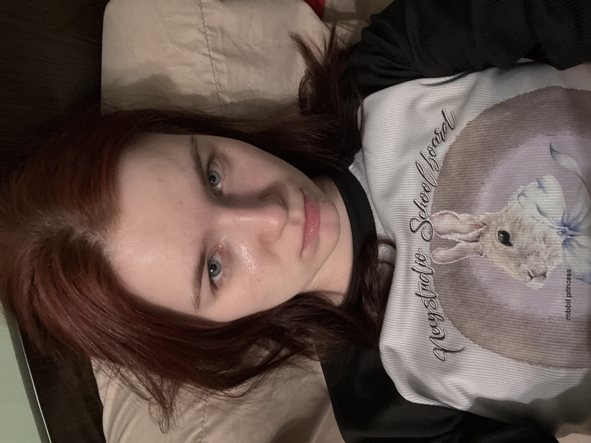 A young woman with auburn hair and blue eyes is lying on a beige pillow, looking directly at the camera with a neutral expression. She is wearing a black and white long-sleeved shirt with a graphic print of a rabbit and text that reads "necy studio school board rabbit princess". The background appears to be a bed with a dark headboard. The image does not provide any information about the city of Sinyavino, Russia.