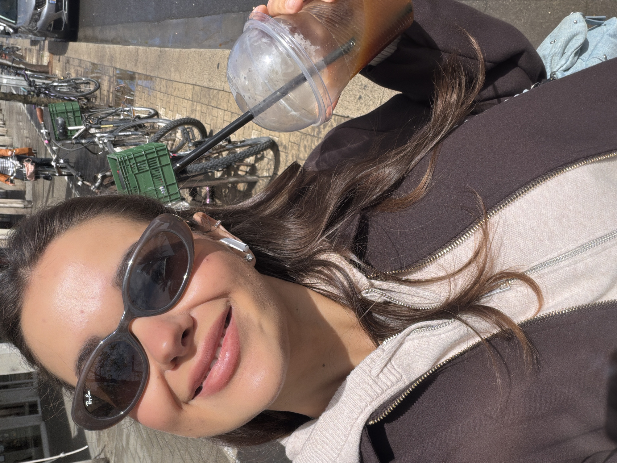 The image captures a selfie-style shot of a young woman enjoying an outdoor, sunny day. The perspective is tilted, with the woman's face appearing on the lower left side of the frame as if she's looking up and to her right, and the background stretching upwards and to the right.
The woman is smiling broadly, revealing her upper teeth. She has smooth skin, lightly tinted lips, and dark eyebrows. Her eyes are concealed by a pair of stylish, dark grey, oval-shaped sunglasses. A "Ray-Ban" logo is visible in white text on the left lens of her sunglasses. Her long, dark brown hair cascades over her shoulders and chest.
She is wearing a dark brown zippered hoodie or jacket over a lighter, possibly beige or off-white, inner zippered garment. Her right hand, partially visible at the top center of the image, is holding a clear plastic cup with a dome lid and a black straw. The cup contains a dark, iced beverage, likely coffee or tea, and condensation is visible on its exterior.
The background suggests an urban environment. A row of bicycles is parked on a paved surface, which appears to be a sidewalk or a street. Several of the bicycles are equipped with bright green plastic crates. Further in the background, a silver car is partially visible on what appears to be a street. The ground surface, possibly brick or cobblestone, shows reflections of sunlight, indicating a bright, sunny day. The overall impression is one of casual enjoyment in an outdoor setting.