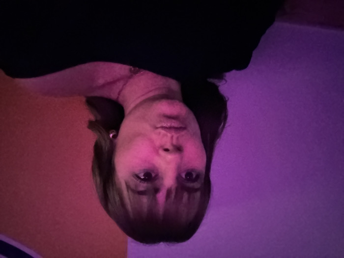 A woman with bangs stares directly at the viewer. Her face is illuminated with a pinkish-red light. She wears a dark top. The background behind her head is divided into two areas; one is a vibrant orange color, and the other is a shade of purple. The image is rotated at 180 degrees. She has an earring on her left ear and is wearing a necklace.