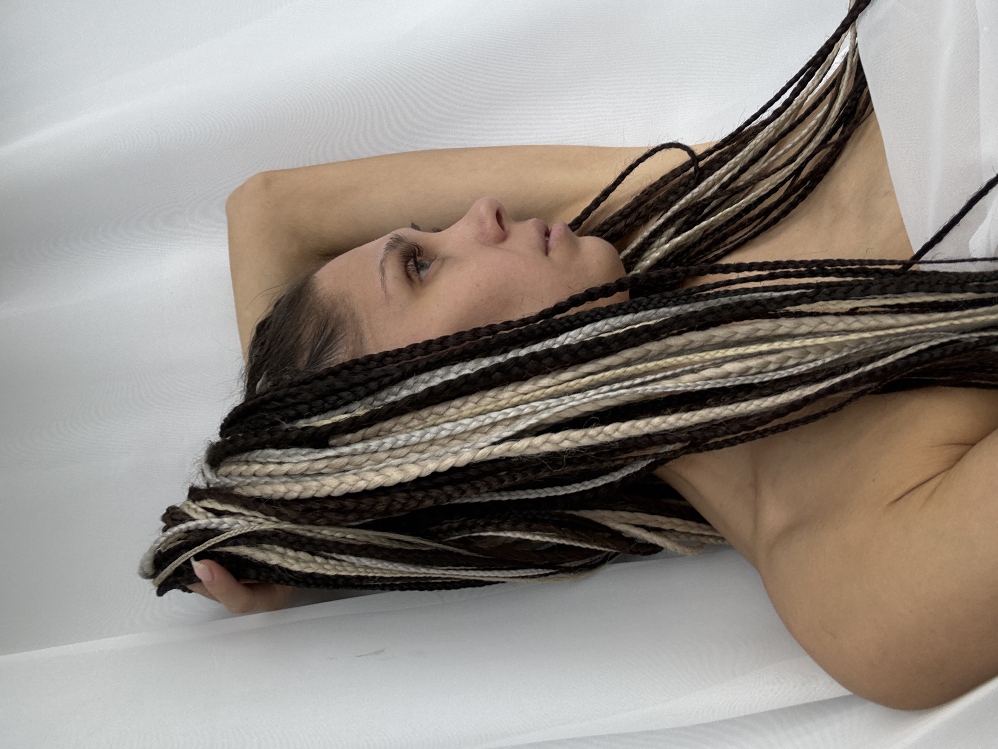 A close-up, horizontal portrait features a person, likely a woman, lying on their back with their head turned to the right. The individual has long, multi-toned braided hair (dreadlocks or similar style) in shades of dark brown, light brown, blonde, and grey, which is spread out across the frame. One arm is visible, bent at the elbow, appearing to support the head or rest behind it, while another bare shoulder is visible on the right. The person's face is in profile, with their eyes open, looking upwards or slightly to the right, conveying a calm or contemplative expression. Their skin tone is smooth and appears to be bare-shouldered. The background consists of a plain, light-colored, possibly white or off-white, fabric that is softly draped, creating gentle folds and textures. The lighting is soft and even, highlighting the subject without harsh shadows. The image captures a serene moment within what appears to be an indoor, controlled environment, potentially in Rusanovo, Russia. There is no visible text in the image.