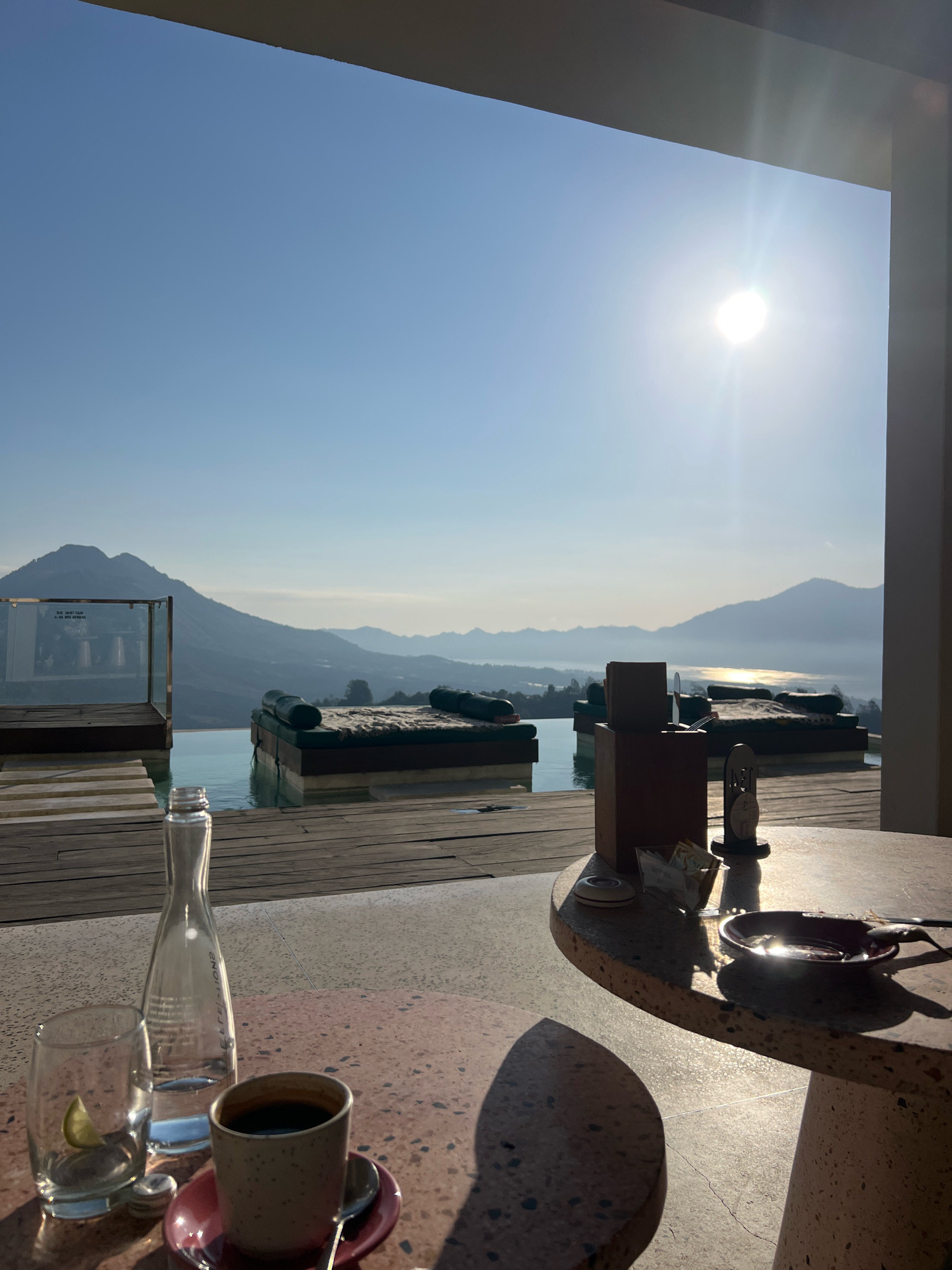 The image shows a serene morning scene in Kintamani, Indonesia, overlooking a vast caldera lake and mountains. In the foreground, a round, speckled table holds a cup of black coffee on a pink saucer, a glass with a lime wedge, and a glass bottle of water. Adjacent to the table, a wooden deck leads to an infinity pool. Several cushioned loungers are positioned around the pool, some partially submerged in the water, offering unobstructed views of the tranquil landscape. The sun shines brightly in the clear blue sky, casting long shadows and reflecting off the calm waters of the lake, creating a picturesque and peaceful atmosphere. The scene is framed by a partial view of a building's overhang, suggesting a resort or cafe setting.
