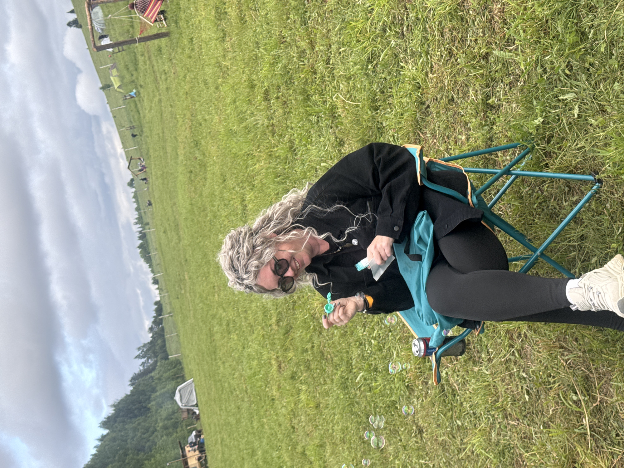 An adult female is seated in a teal and orange foldable camping chair on a grassy field in Большая Кивара, Russia. She has wavy, light-colored hair, wears dark sunglasses, a black long-sleeved garment, black leggings, and light-colored athletic shoes. Her right hand holds a green bubble wand, and her left hand holds a small clear pouch. Several iridescent bubbles are visible floating to her left. A red and silver beverage can is secured in a mesh holder attached to the chair's right armrest. The background consists of an ascending green field under a predominantly cloudy sky. To the left, a dense forest covers a hillside, with a light-colored tent, a wooden structure, and a dark vehicle situated at the base of the incline. To the right, the field continues upwards, featuring multiple wooden posts, several additional small tents, and a prominent wooden swing set with a person, partially obscured by a striped covering, seated within.