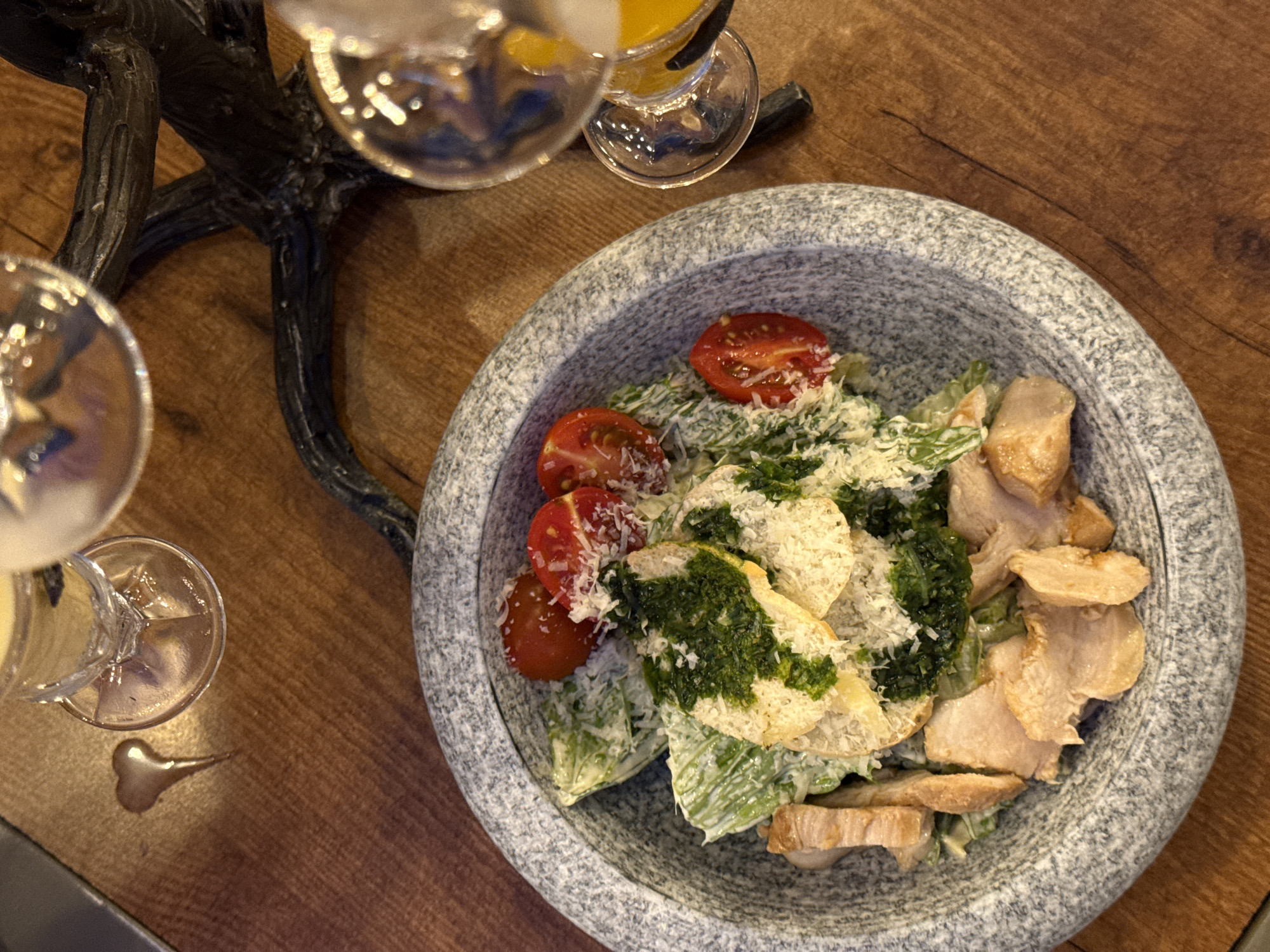 The image depicts a plate of Caesar salad on a wooden table. The salad is composed of lettuce, sliced tomatoes, croutons with green pesto, and chicken. The salad is topped with grated Parmesan cheese and served in a large gray stone bowl. There are two glasses with a light-colored drink (likely cocktails) near the salad bowl. The location is Kazan, Russia.