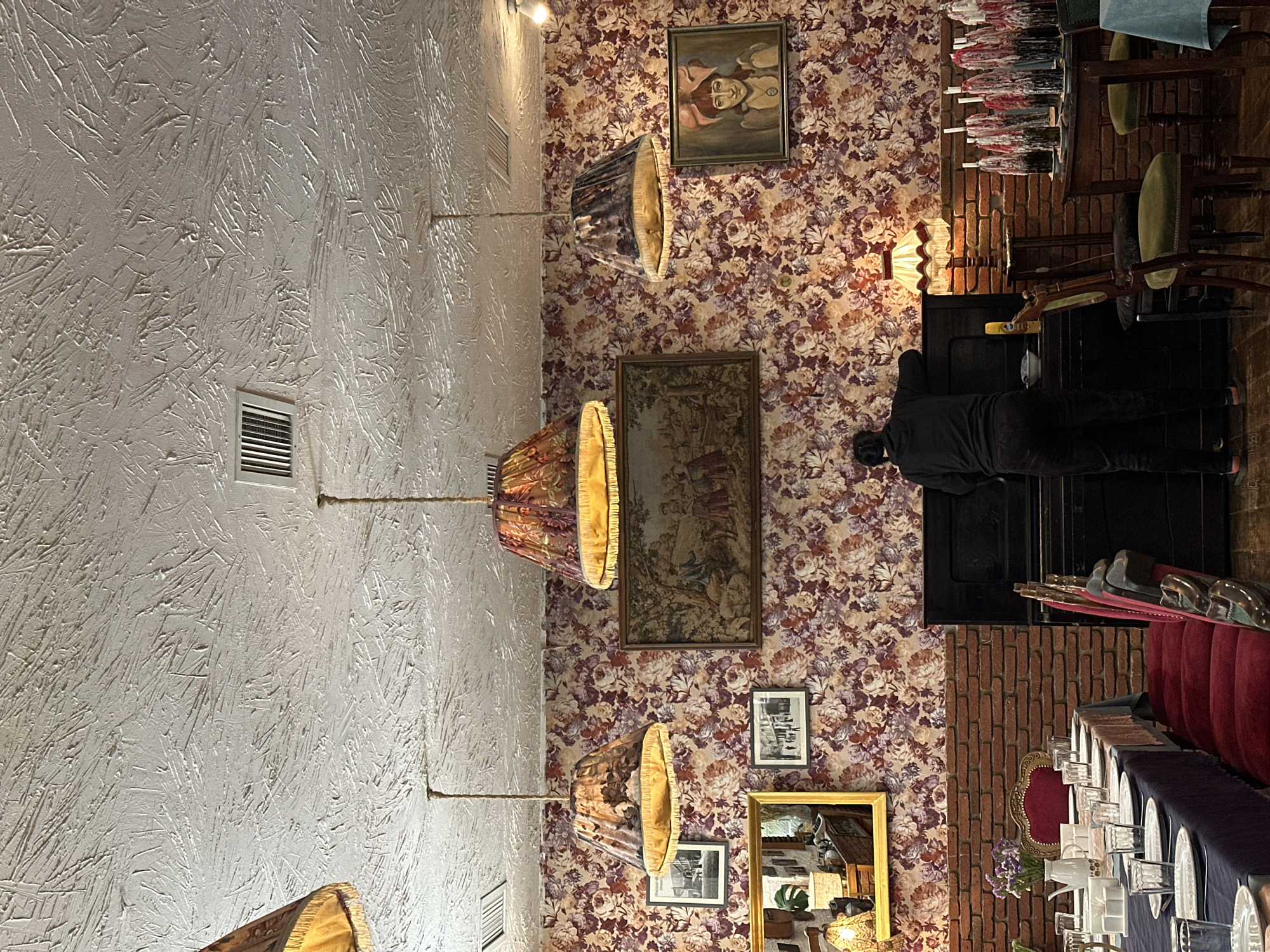 The image shows the interior of a restaurant with eclectic décor. The walls are covered in floral wallpaper with a white textured wall on the left side. There are framed pictures and a mirror hanging on the floral wallpapered walls. There are several lamps with ornate lampshades. In the background, there is a person dressed in black standing near a fireplace. To the right, there is a long table set for a formal dinner, with glassware, plates, and cutlery, covered in a blue tablecloth with red upholstered chairs. The location is likely Tbilisi, Georgia, given the architecture and decorative elements.