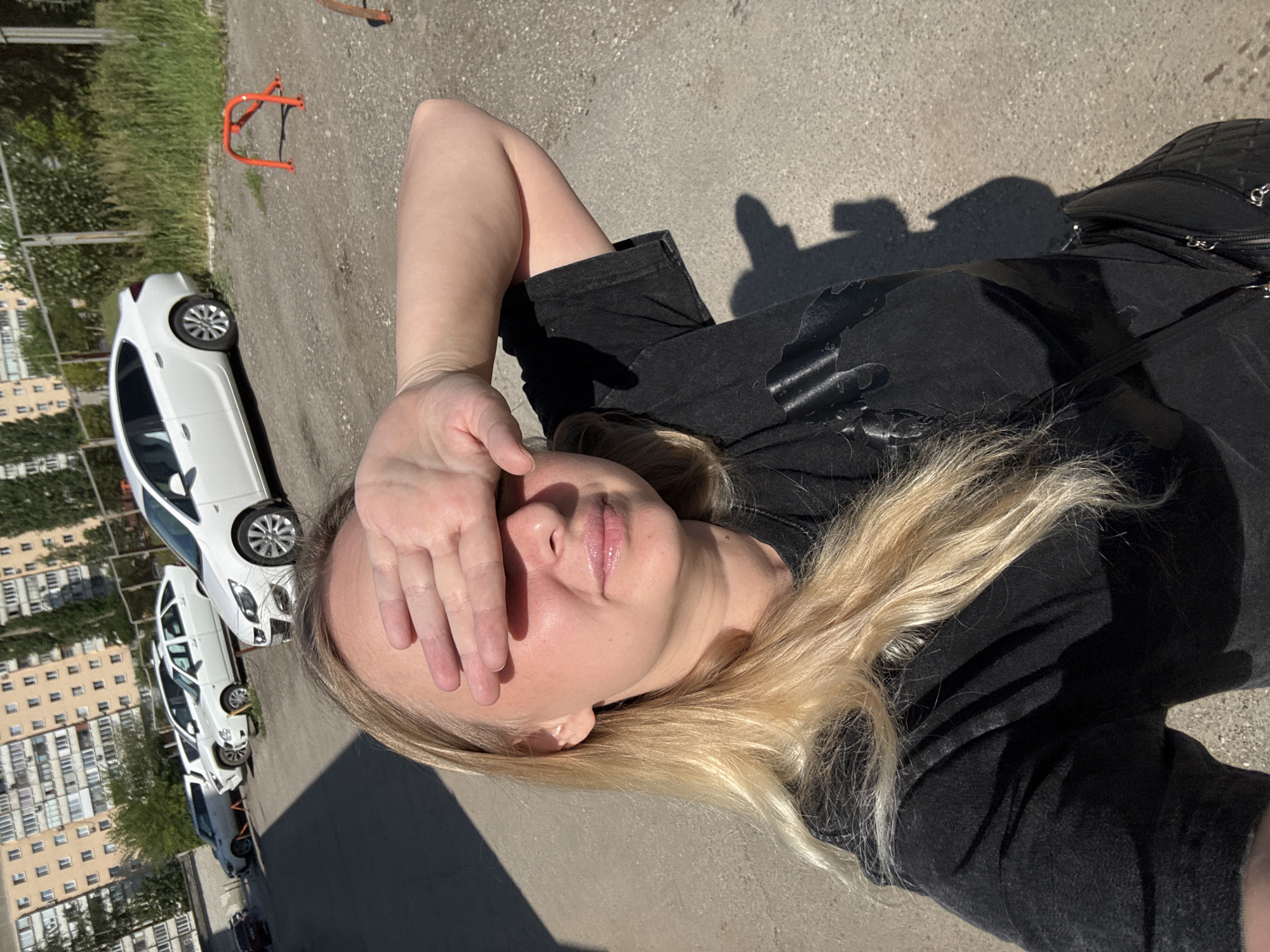 A blonde woman is taking a selfie outdoors in Saratov, Russia. She is wearing a black t-shirt and has her right hand covering her eyes. Behind her, several white cars are parked in a lot, and a multi-story apartment building is visible in the background. The ground is covered in gravel and pavement. There is a metal barrier painted orange in the middle ground.