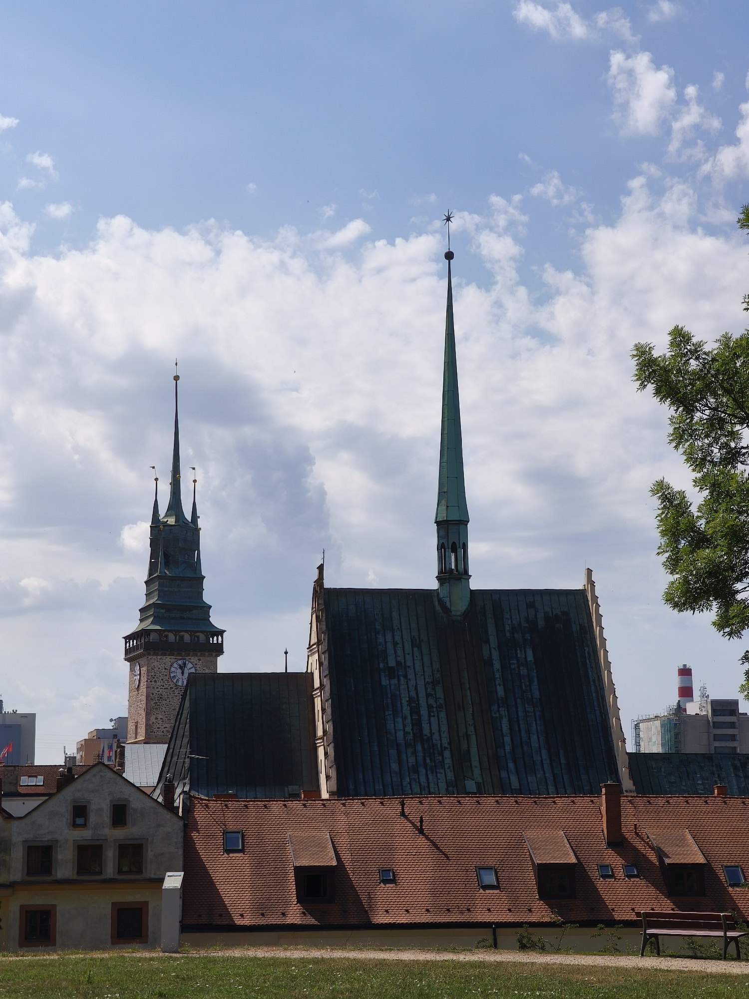 The image shows a city skyline dominated by several prominent buildings, including two church spires and a clock tower. In the foreground, a low-rise building with a red tile roof and dormer windows is visible, along with a wooden bench on a grassy area. In the background, modern buildings and industrial structures, including a tall tower with red and white bands, are visible. The sky is blue with scattered white clouds. The scene is a daytime view of the city. The church spires and clock tower indicate a historic part of the city, while the modern buildings suggest urban development. This photograph was taken in Pardubice, Czechia, showcasing the juxtaposition of historical architecture and modern infrastructure. The perspective is from an elevated position looking towards the city.