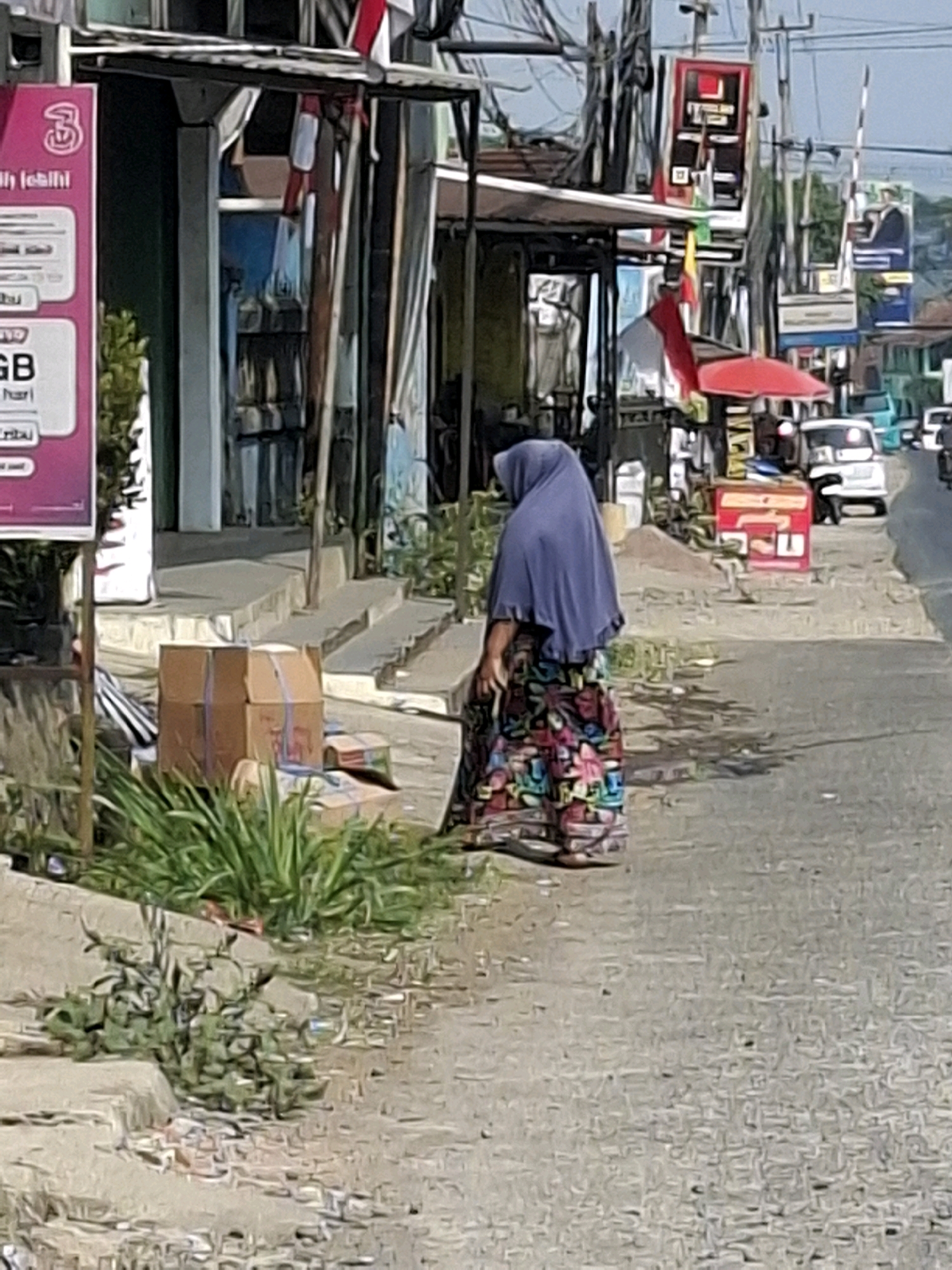 A full shot captures a street scene in Lembursawah, Indonesia, under a bright sky. In the foreground, a woman wearing a blue hijab and a colorful floral skirt walks away from the camera. She is on the right side of the image, with the street stretching out before her. To the left, a store with a red and white sign stands beside a stack of cardboard boxes and lush green plants. The store's entrance is visible, leading into a dark interior. The street is lined with various buildings and signs, and several vehicles are parked or driving in the background, indicating an active urban environment. The overall scene is illuminated by natural light, suggesting it is daytime.