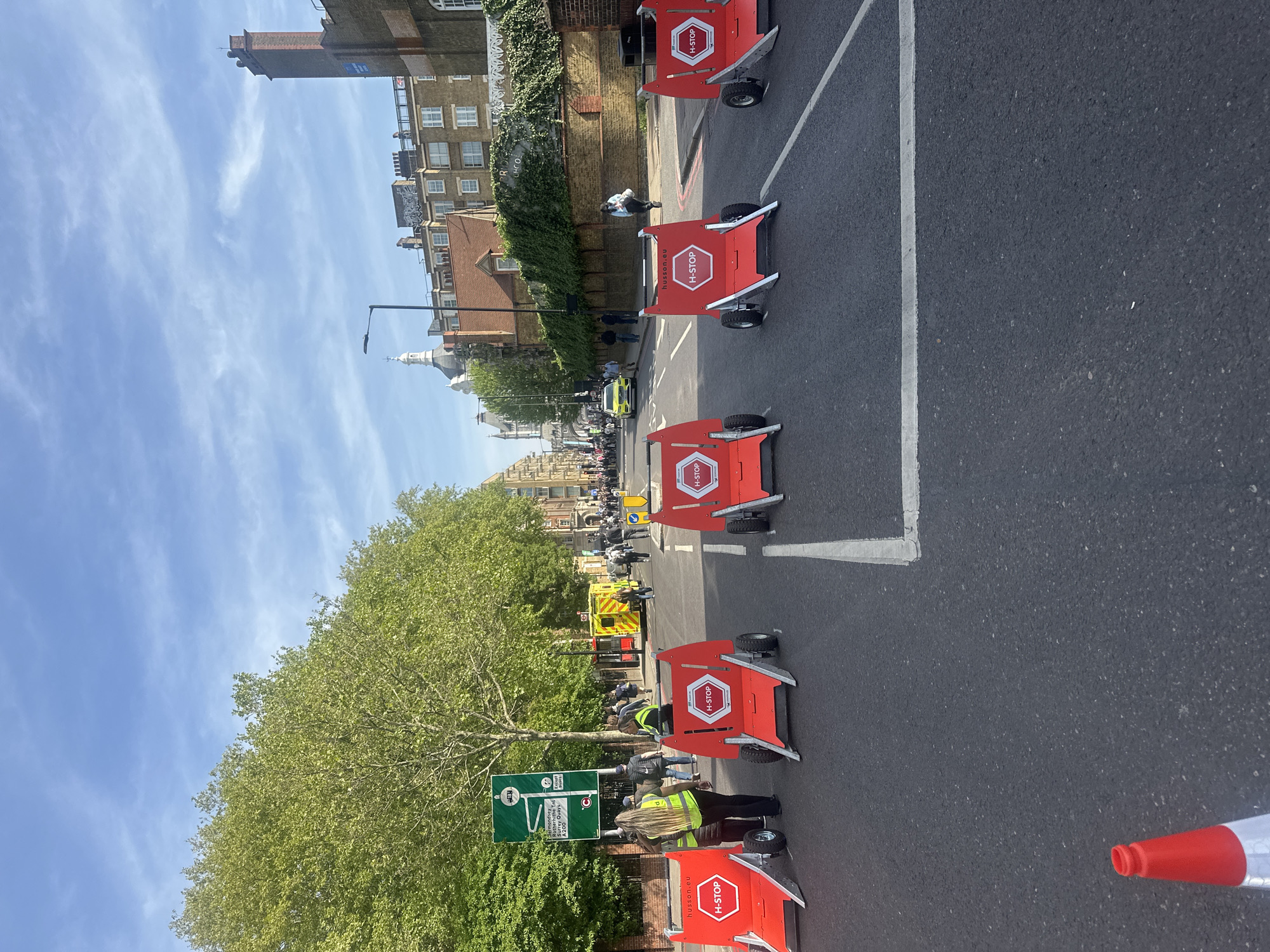 The image captures a street scene in London, United Kingdom. The road is lined with red and white H-STOP barricades, indicating a potential closure or restricted area. Several individuals are present, some wearing high-visibility vests, suggesting they may be workers or event staff. In the background, there are buildings, including a notable one with a dome, common in London's architectural landscape. Street signs are visible, suggesting routes to Rotherhithe, the A200, and Surrey Quays. Additionally, an ambulance is observed further down the road, possibly related to the activities occurring on the street. The overall environment suggests a planned event, road work, or situation managed by safety personnel.
