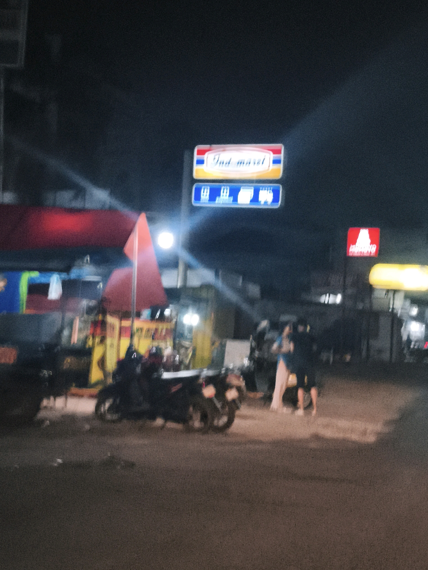 The image depicts a nighttime street scene in Palembang, Indonesia, likely in an urban or commercial area. In the background, a sign for "Indomaret," a popular Indonesian convenience store chain, is prominently displayed with its characteristic red and yellow logo. Below the Indomaret sign is a blue sign with white symbols, possibly indicating services or directions. To the right, another red sign with white lettering, possibly another commercial establishment, is visible.
The street is paved and appears wet, suggesting recent rain or street cleaning. Several motorcycles are parked or in motion on the street, some with riders. In the middle ground, there are structures that appear to be market stalls or small shops, some adorned with colorful fabrics and lighting. A bright white light source, possibly a streetlamp or a strong light from a shop, illuminates part of the scene.
In the foreground, a person wearing a blue top and a light-colored skirt or dress is interacting with another individual who is partially obscured. Their body language suggests a casual conversation or exchange. The overall atmosphere is that of a bustling commercial area at night, with the glow of signs and lights illuminating the street and its activities.
