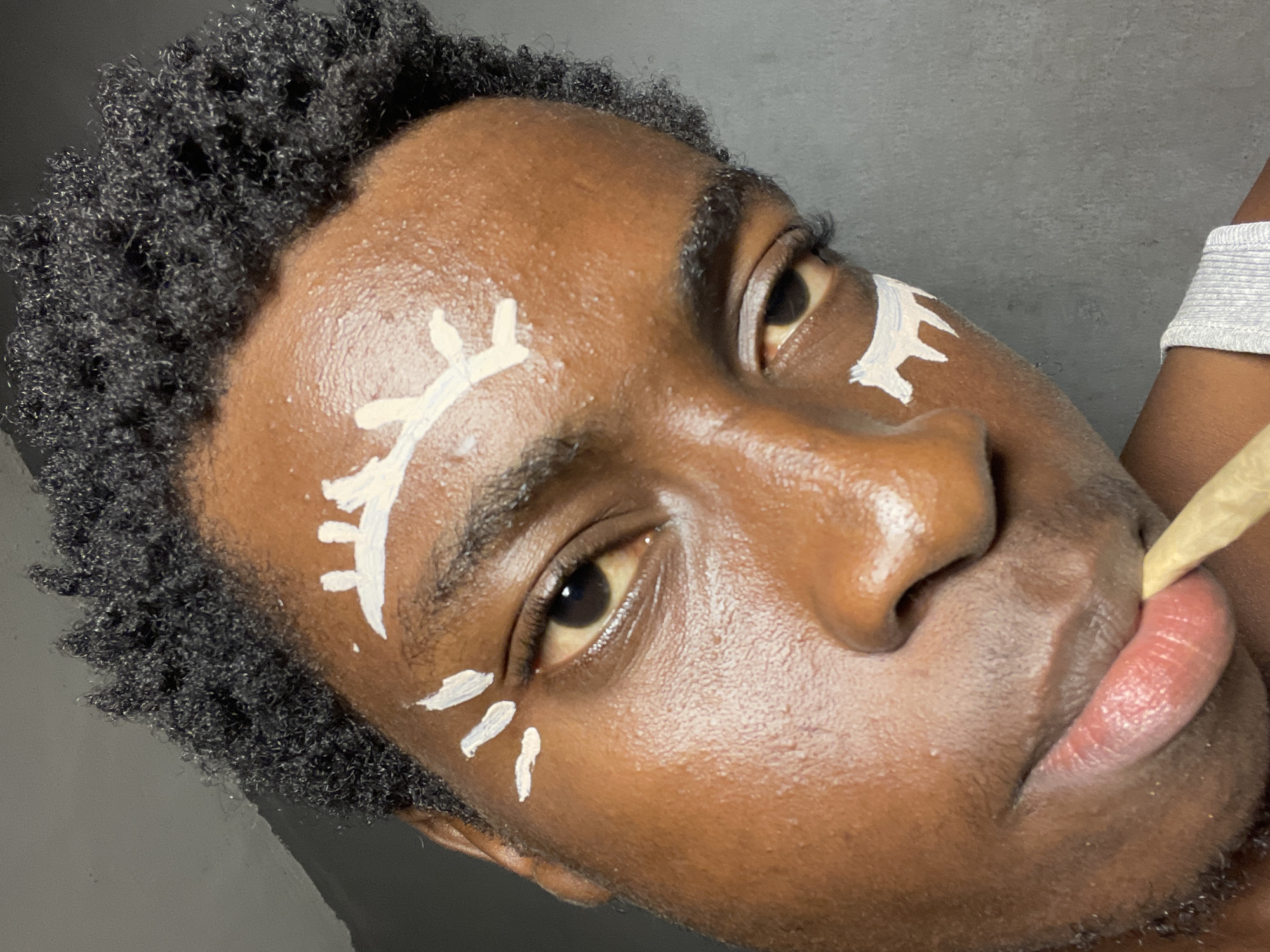 The media file is a close-up, low-angle shot of a young Black person's face, angled upwards and to the left. The individual has dark, curly hair and is wearing white face paint in abstract designs on their forehead and below their eye. The skin appears dewy, and the lighting suggests an indoor setting. The person's mouth is slightly open, with what appears to be a rolled cigarette or joint held between their lips. The background is a plain, dark gray wall. The overall mood is one of artistic expression or personal adornment. The time of day and weather are not discernible, and there are no visible text elements. The location context provided is Nkpor, Nigeria.