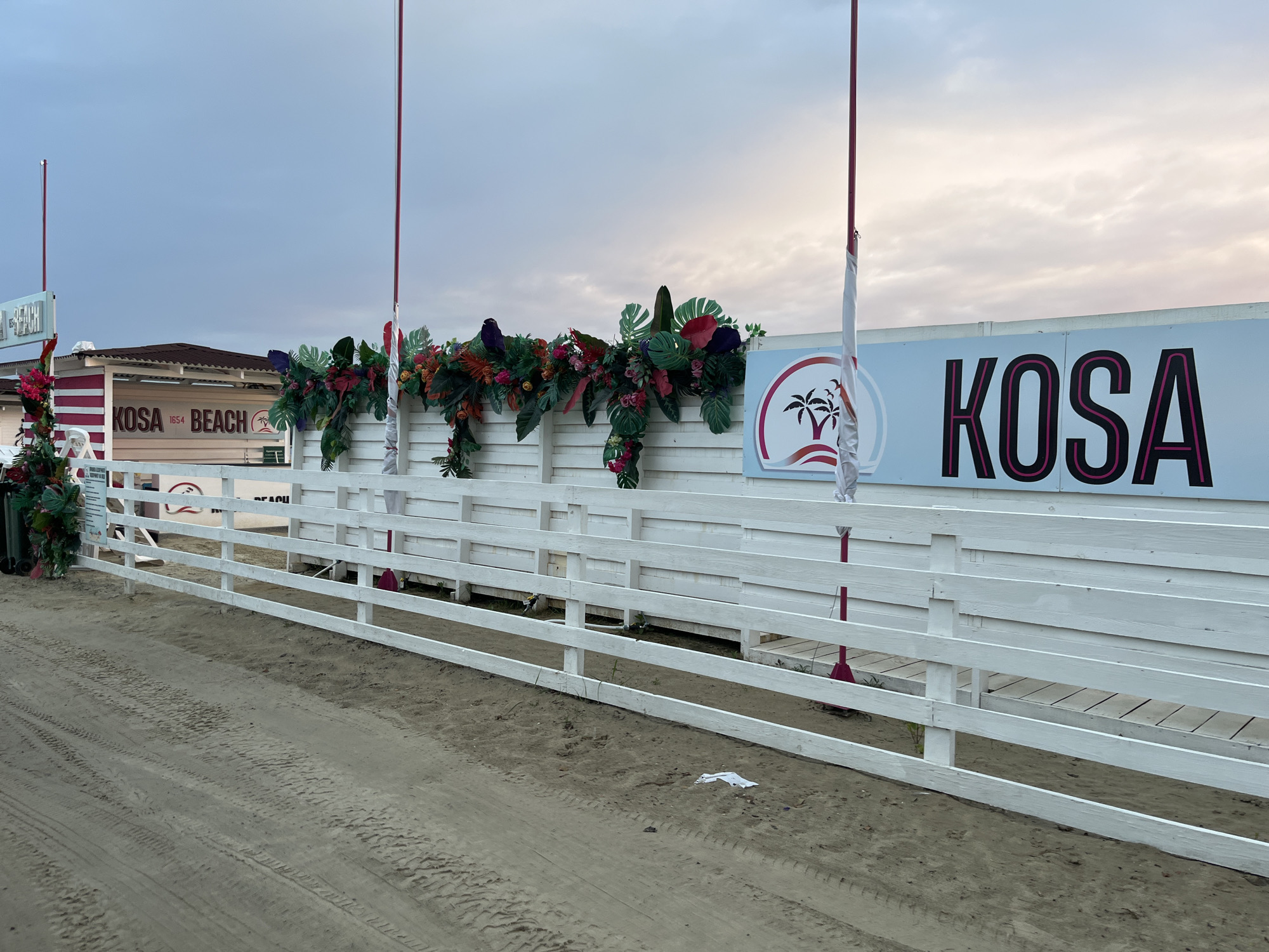 The image depicts an outdoor scene at KOSA BEACH in Bezliudivka, Ukraine. The foreground shows a sandy area with tire tracks. A white wooden fence runs horizontally across the middle ground, separating the sandy area from a raised boardwalk further back. Along the top of the fence, there is a decorative garland of artificial tropical leaves and flowers. Behind the fence and garland, a building with "KOSA BEACH 1654" written on it is visible, along with a large blue sign featuring the word "KOSA" in bold pink lettering and a logo with palm trees. Two red flagpoles with white flags attached are also present. The sky is overcast with hints of pink and orange light, suggesting either sunrise or sunset.