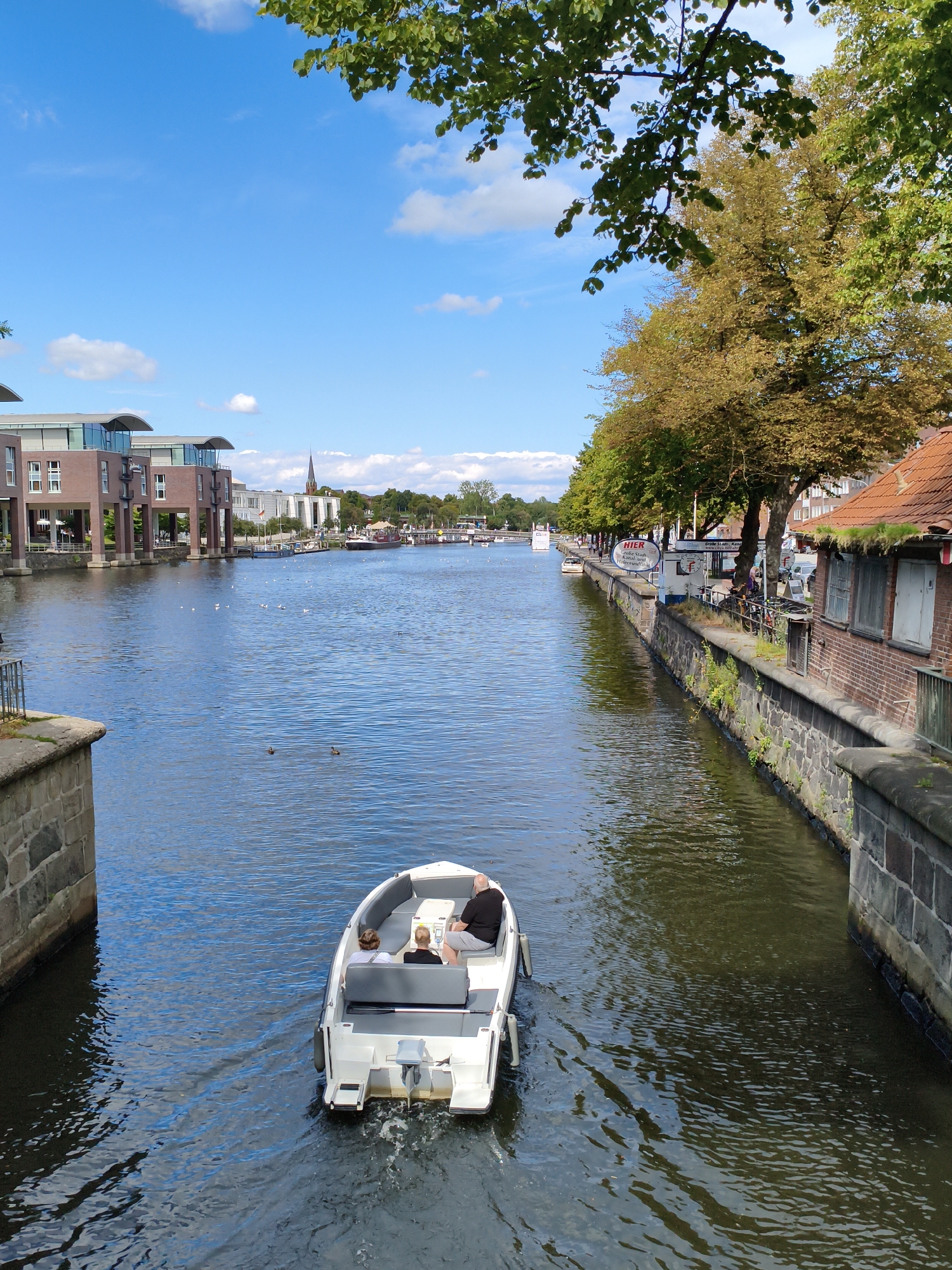A bright, sunny day unfolds over a calm, blue waterway in Lübeck, Germany, as a small white motorboat carrying three passengers navigates through the canal. The boat is positioned in the foreground, moving away from the viewer, with two individuals seated in the back and one in the front. On the left bank of the canal, a modern brick building with large glass windows stands, reflecting the sky. On the right bank, a traditional brick building with a red tiled roof and weathered windows is visible, with trees lining the canal. In the distance, a church spire pierces the sky, and other boats are moored along the waterfront, suggesting a bustling urban environment. The water's surface is slightly rippled, and several ducks can be seen swimming peacefully. The scene is framed by lush green foliage from trees overhead, casting dappled shadows on the water.
