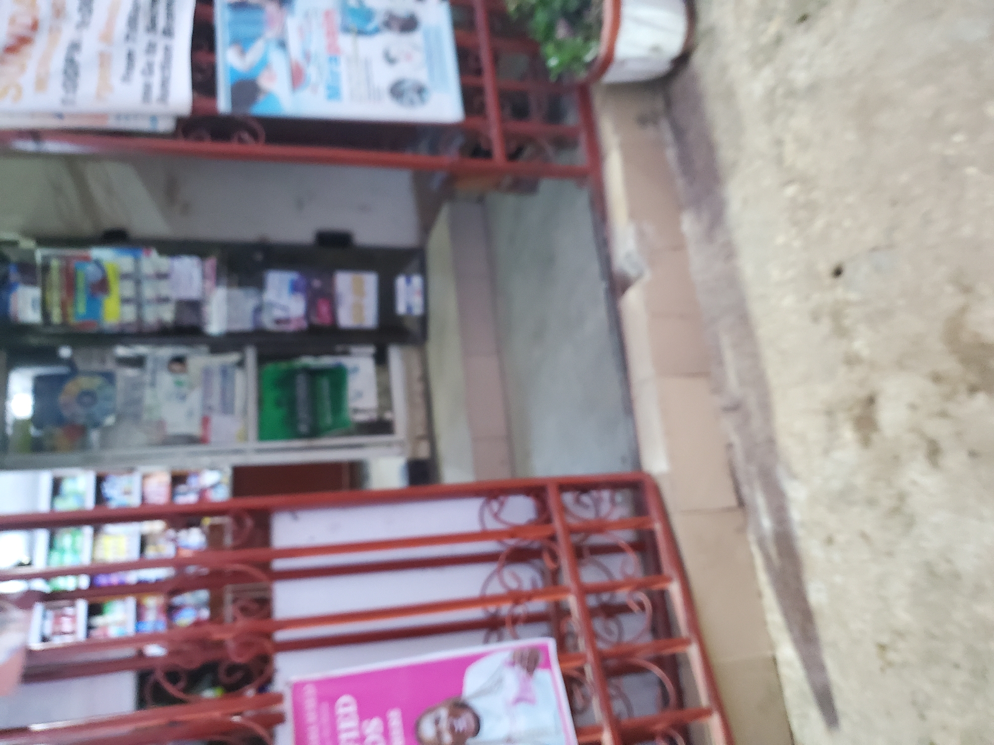 The image depicts a blurry outdoor scene in front of a store, possibly a pharmacy or convenience store, located in Jos, Nigeria. A red metal gate is partially open, revealing shelves stocked with various items inside the store. Posters are affixed to the exterior walls. One poster on the left features a person's image and text, while another on the right has text and graphics related to a product or service. A white potted plant is visible to the right of the gate. The ground in front of the store appears to be paved with concrete or rough asphalt. The overall impression is of a commercial establishment in a public area.