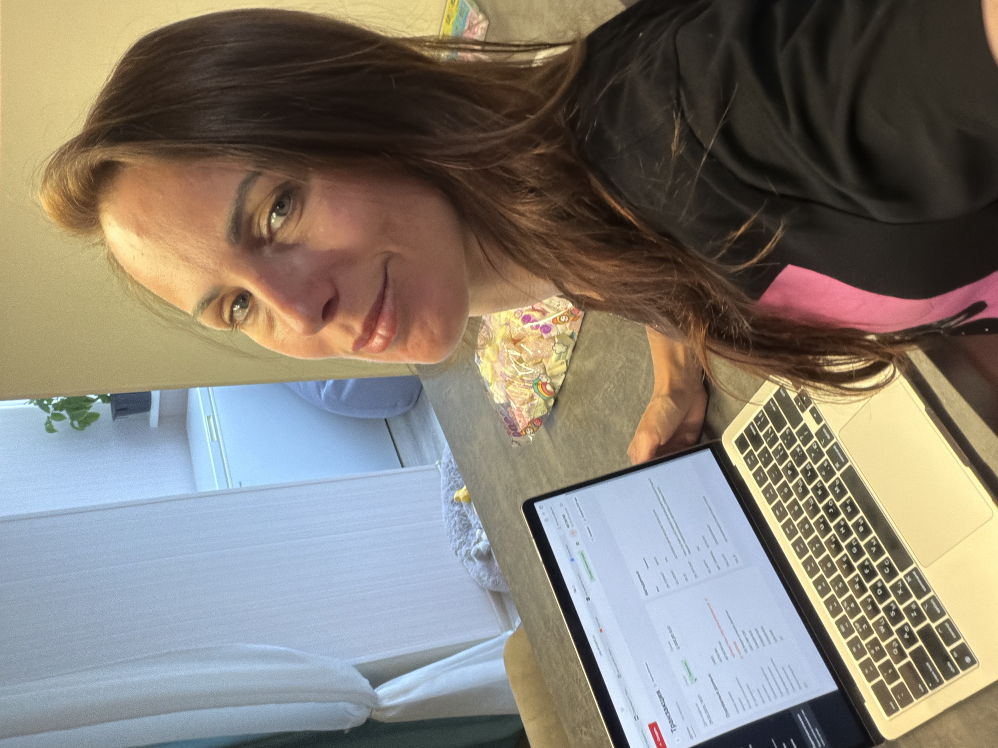The image features a woman indoors, looking directly at the camera with a gentle smile. She has long brown hair and is wearing a black top with a bright pink accent visible on the lower left side. Her right arm and hand are positioned near a silver laptop, which is open on a flat, grey surface.
The laptop screen displays what appears to be a web application or software interface, showing structured data in columns, likely a table or form. Visible text on the screen includes Cyrillic characters, such as "Транзакция" (Transaction), along with numerical data. The laptop's keyboard also clearly shows a Cyrillic layout, confirming the use of the Russian language.
In the background, a light-colored wall is visible, and to the woman's left, there's a window covered by white blinds or curtains. A potted plant with green leaves can be seen in the upper left corner of the background. On the surface next to the laptop, a colorful plastic bag with a unicorn pattern and a light-colored, fluffy object, possibly a slipper, are present. The setting suggests a casual indoor environment, likely a home or personal workspace. The lighting indicates it is daytime. The woman's expression conveys a calm and friendly demeanor. The location context for this media file is Birobidzhan, Russia.