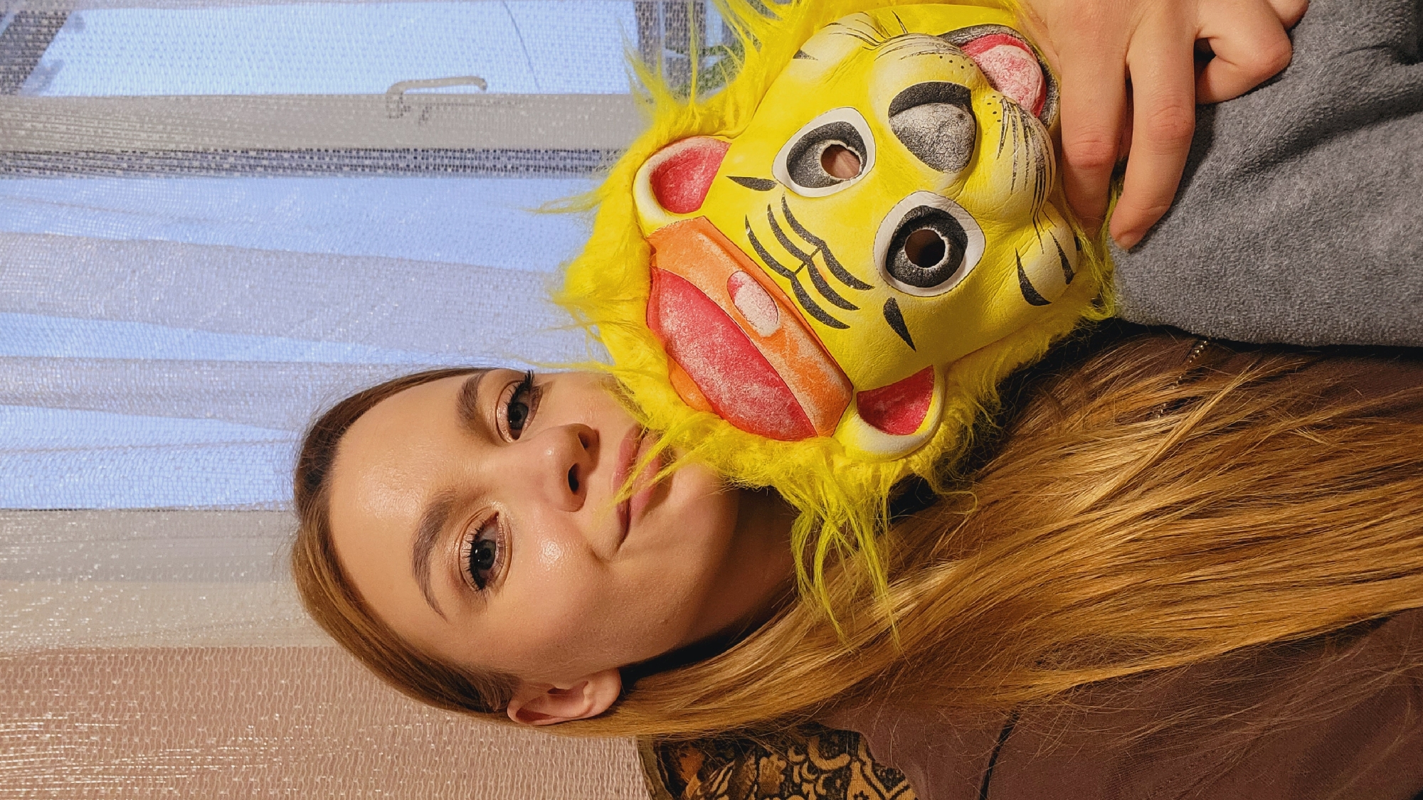 A young woman with long, light brown hair is lying down indoors, holding a bright yellow lion mask to her face. Her head is positioned at the lower left of the frame. The mask is held by a hand visible at the upper right. A window with sheer curtains is visible in the background. The location is assumed to be Зипуново, Russia based on the prompt request, but no specific detail in the image confirms this location.