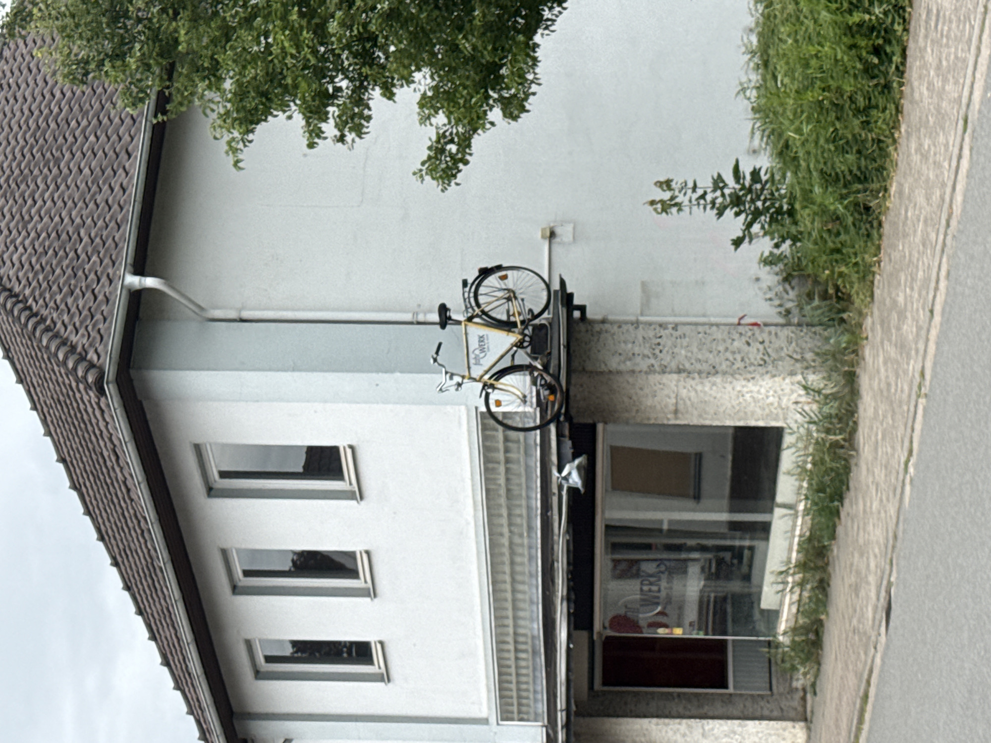 This image captures a building facade in Bassum, Germany. The building has a white upper story with multiple rectangular windows and a dark brown tiled roof. A yellow bicycle is mounted on a metal rack attached to the building's exterior wall, above what appears to be a storefront or entrance with large glass windows. The bicycle is positioned vertically. Green foliage, including tree branches and bushes, frames the scene on the left and right sides. The ground in front of the building is paved with light-colored concrete slabs. A sidewalk runs alongside the building. The overall impression is one of an urban or semi-urban setting. The lighting suggests it could be daytime, with overcast skies indicated by the soft, diffused light. There is some visible text on the building's glass facade, including the word "WERK" which is likely part of a business name. The image is oriented vertically.