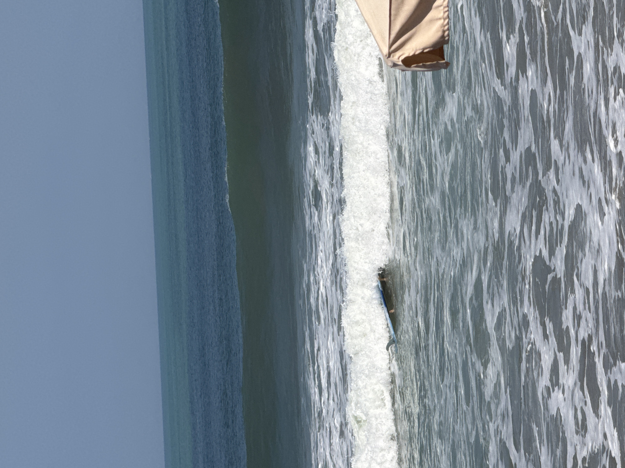 The image depicts a beach scene in Kuta, Indonesia, with the ocean occupying the majority of the frame. The water is a mix of deep blue and turquoise hues, with several layers of waves visible. The most prominent feature in the water is a surfboard, partially submerged and angled towards the left side of the image. A person appears to be lying on the surfboard, or perhaps has fallen off and is in the process of getting back on. Their legs are visible, dangling in the water near the back of the board. The surfboard has a fin clearly visible at its tail. In the foreground, the bottom right corner of a tan beach umbrella is visible, suggesting a populated beach area. The sky above is a clear, pale blue.