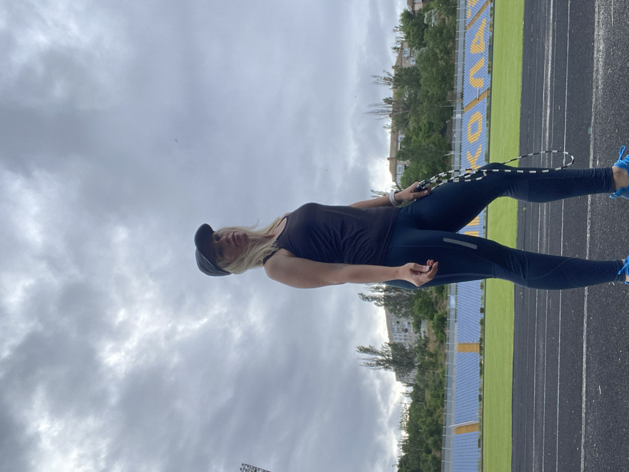 An outdoor, low-angle, full shot shows a woman in athletic wear standing on a running track in front of a stadium. She is wearing a black tank top, dark blue leggings, a dark blue baseball cap, and bright blue athletic shoes. She holds a black and white beaded jump rope in her right hand. Her left hand is by her side. The stadium seating is blue and yellow, and a green field is visible behind the track. The sky is overcast with gray clouds. Trees and buildings are in the background.