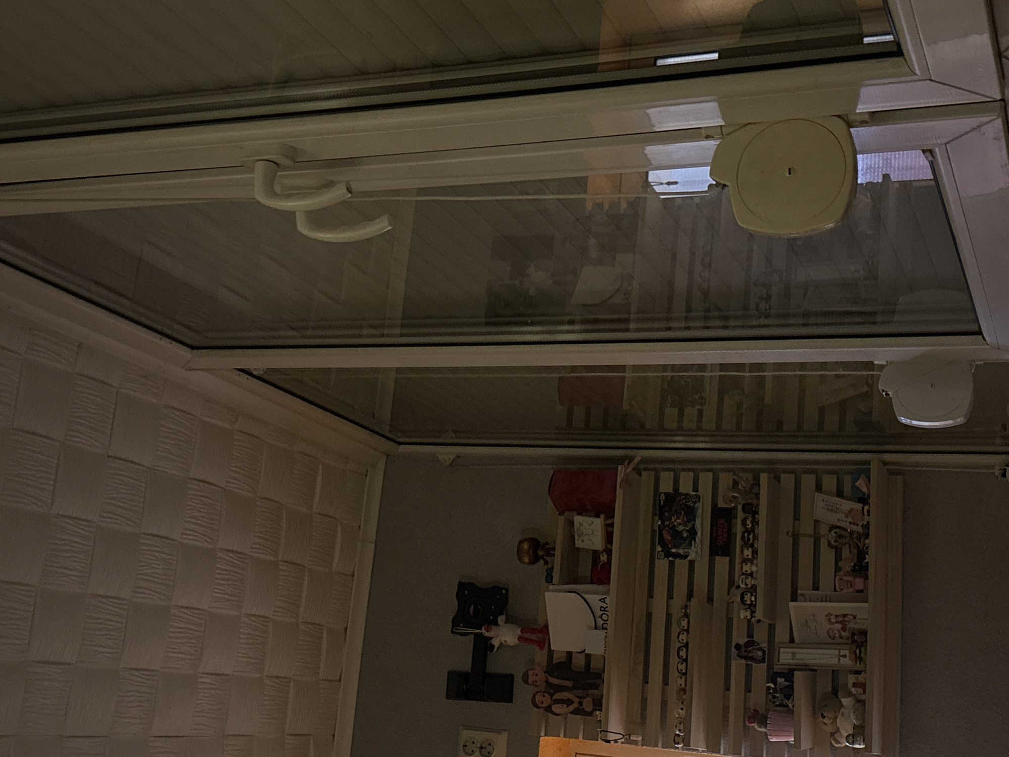 The image shows an interior view of a room, likely in Panagyurishte, Bulgaria, as requested. The perspective is tilted, looking upwards at a white-framed window or skylight. The window has a handle on the left side and a round, white mechanism (possibly for blinds or a shade) on the right. Through the glass, a reflection of the room's contents can be seen. Beneath the window, a wall with textured, woven paneling is visible on the left. On the right, a collection of objects is displayed on a wooden shelf unit. The objects include figurines, pictures, and boxes; one box has the word "PANDORA" visible. Other items like a wall-mounted TV bracket and an electrical outlet are also in view. The lighting is dim. There are no people visible in the image. The focus is mainly on the architectural details and reflected objects.