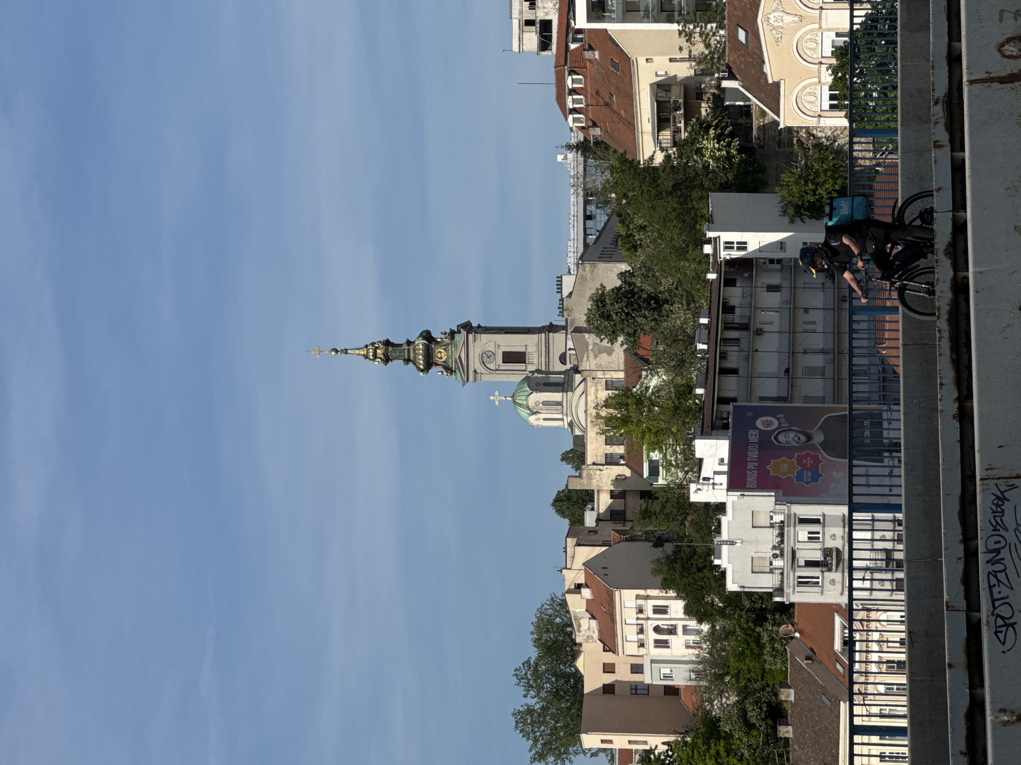 A street scene in Belgrade, Serbia is viewed from an elevated position, possibly a bridge. The shot is aligned vertically with the sky occupying the left side of the frame. The architectural structure of the Church of Saint Alexander Nevsky is visible, with its elongated spire rising towards the sky. To the right of the church, various buildings, typical of an urban landscape, are seen, some with trees interspersed among them. A person on a bicycle, wearing a dark outfit and carrying a blue delivery bag, rides along the bridge to the right side of the frame. A large advertisement is visible on one of the buildings, and graffiti marks the structure from which the photo is taken. The lighting suggests it is daytime with clear skies.