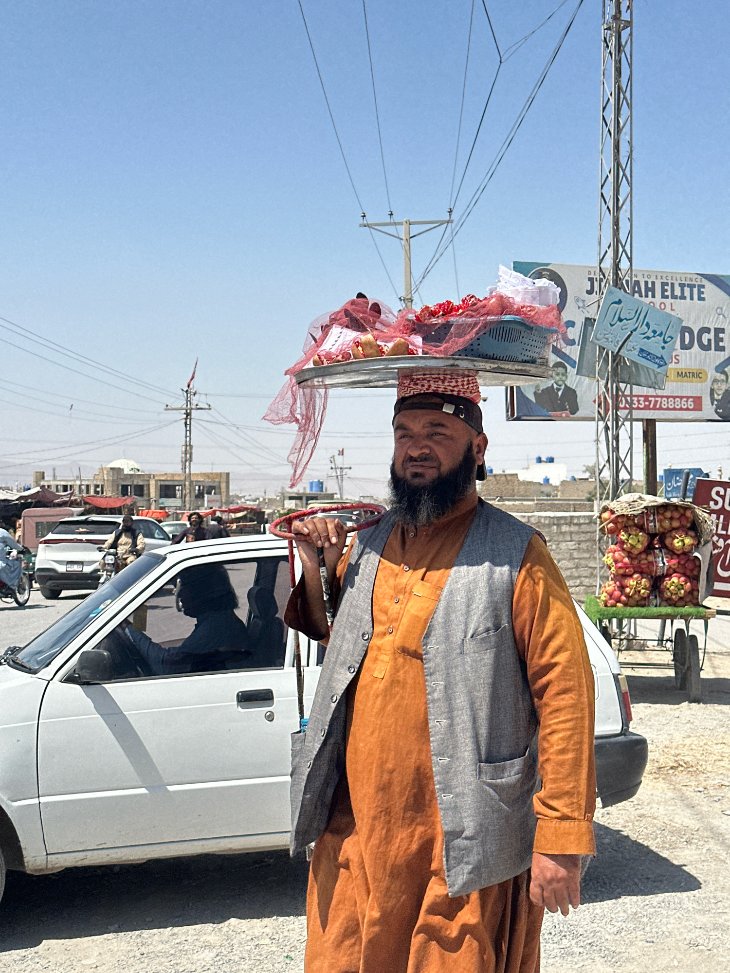 A man with a black beard and wearing a traditional shalwar kameez and vest walks down a street in Quetta City Tehsil, Pakistan. He carries a tray of goods on his head. The tray contains a blue basket filled with items wrapped in red netting. To his right, a cart is laden with red apples. The man is walking alongside a white car. In the background, there are buildings, power lines, and a billboard. The sky is clear and blue, indicating a sunny day. The ground is unpaved and appears to be a mix of dirt and gravel.